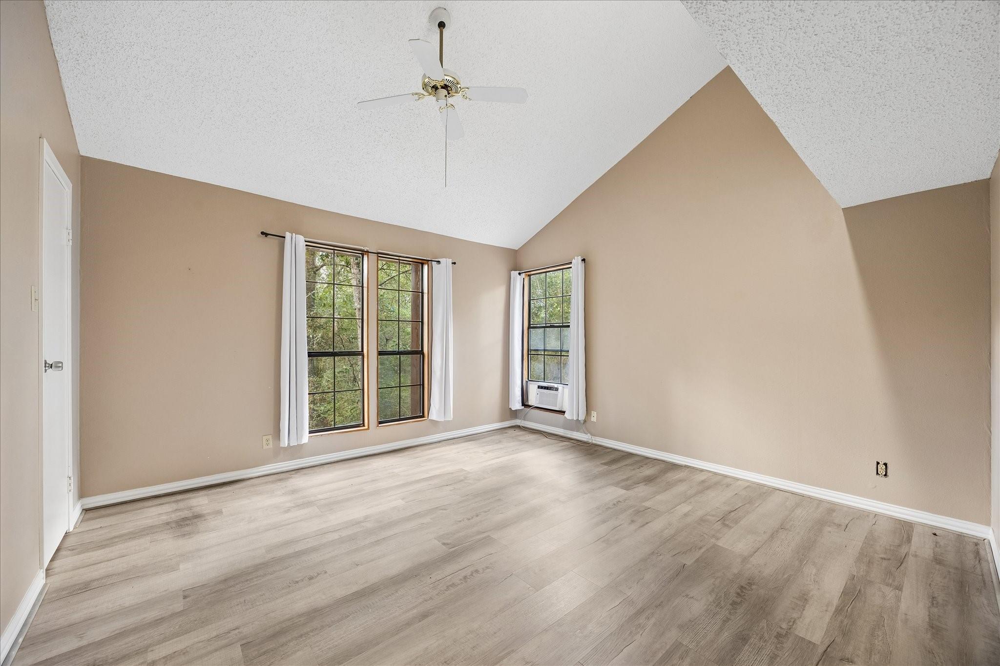 Vacant bedroom with vaulted ceiling, light wood-look floors, curtained windows and a window air conditioner.