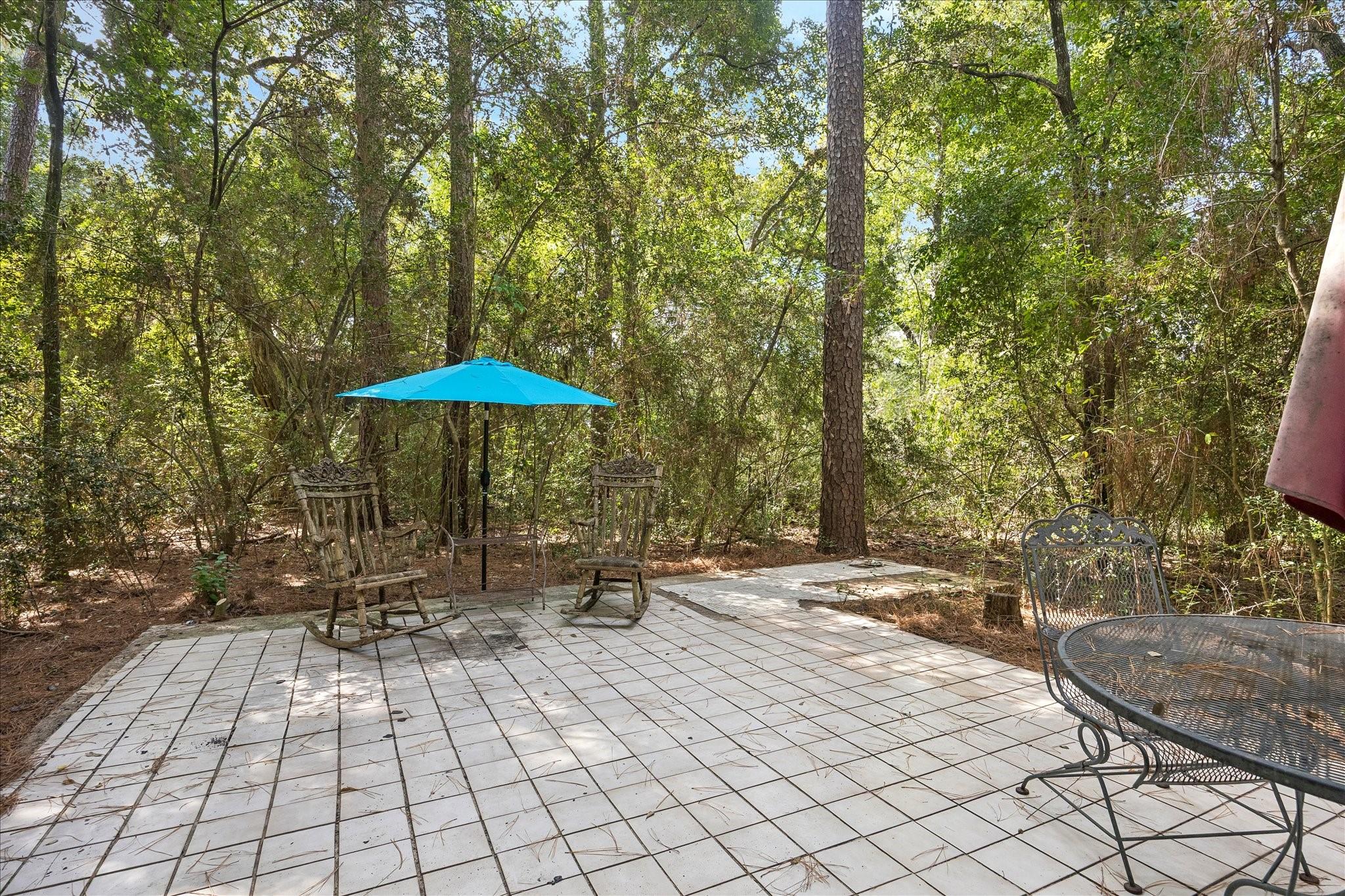 Tiled patio with outdoor chairs and a blue umbrella surrounded by dense trees.
