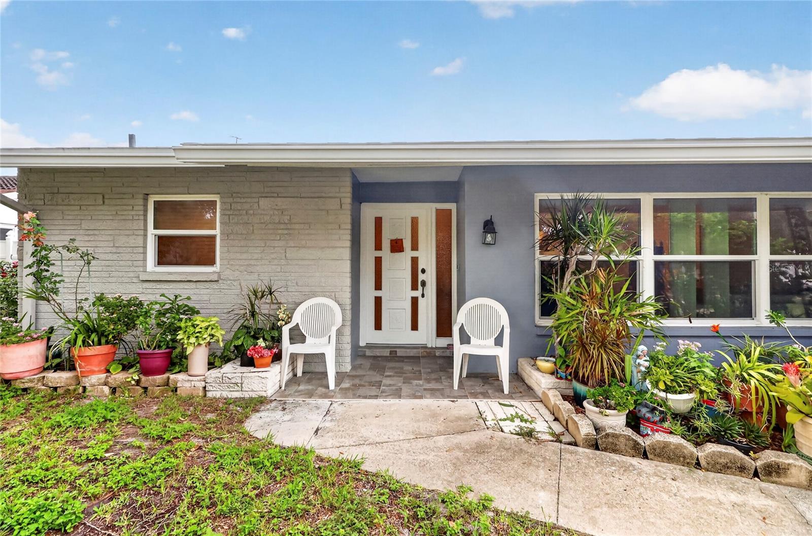 Single-story home with a recessed white entry, two chairs, and colorful plants along the walkway.