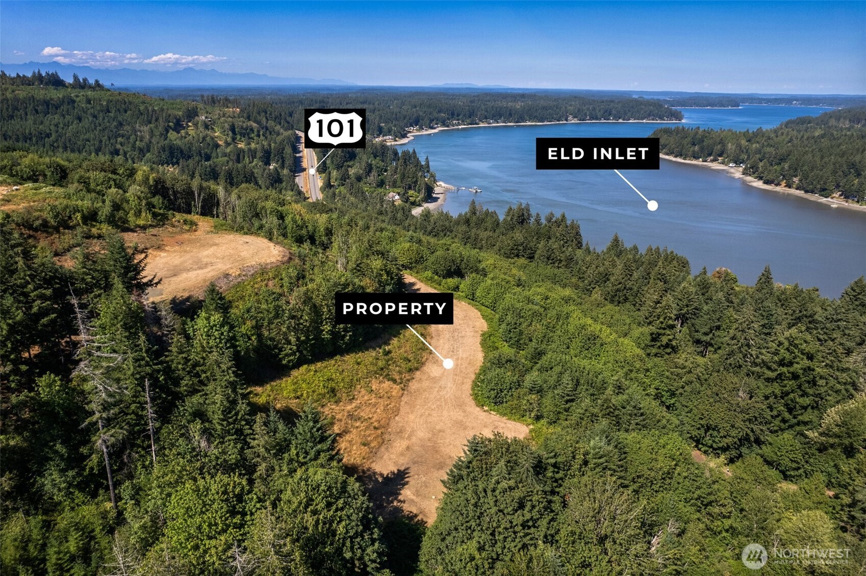 Cleared property surrounded by forest beside Eld Inlet, with Highway 101 and distant mountains visible.