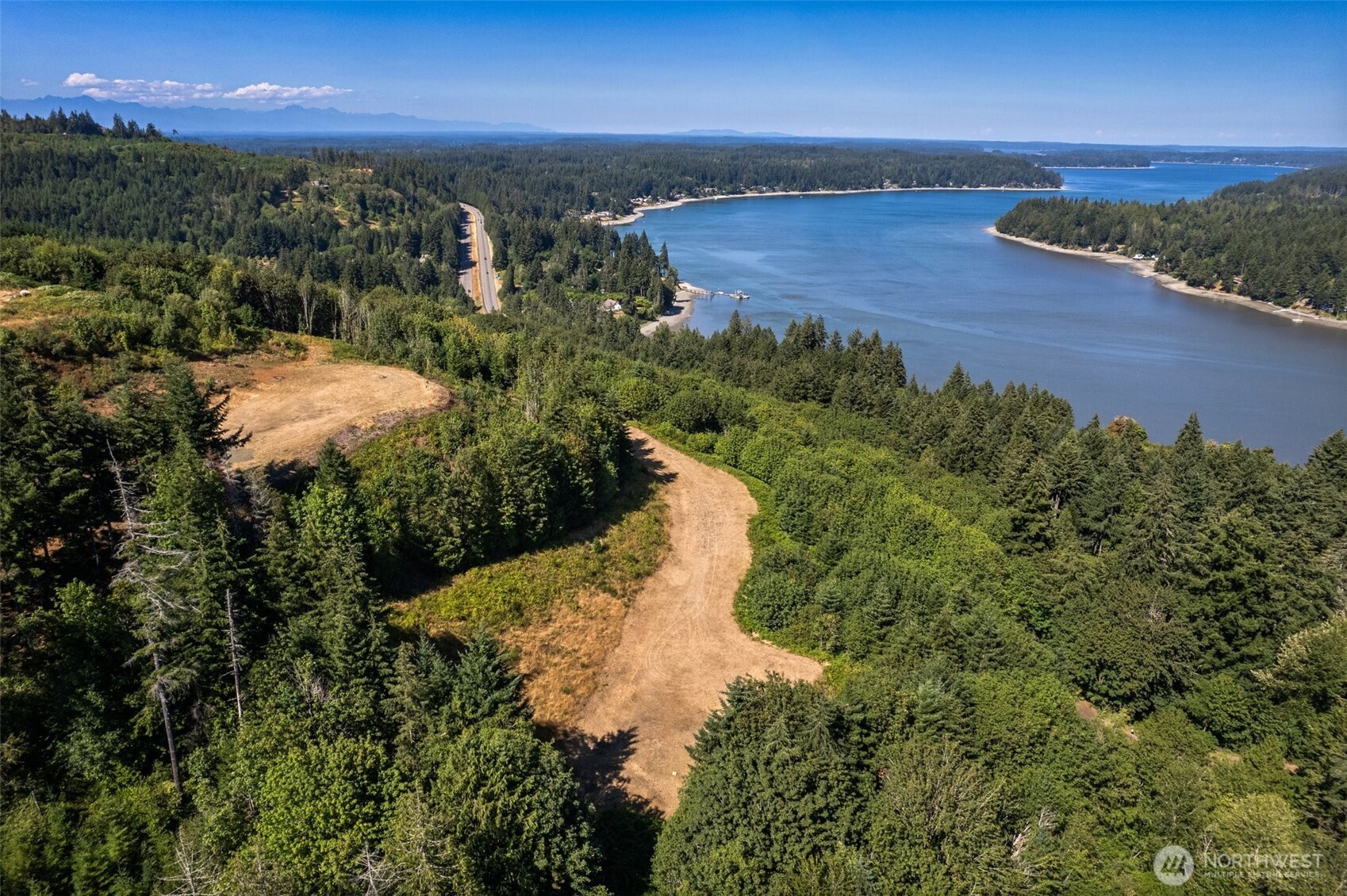 Aerial view of a wooded clearing beside an inlet, with forested shores and distant mountains.