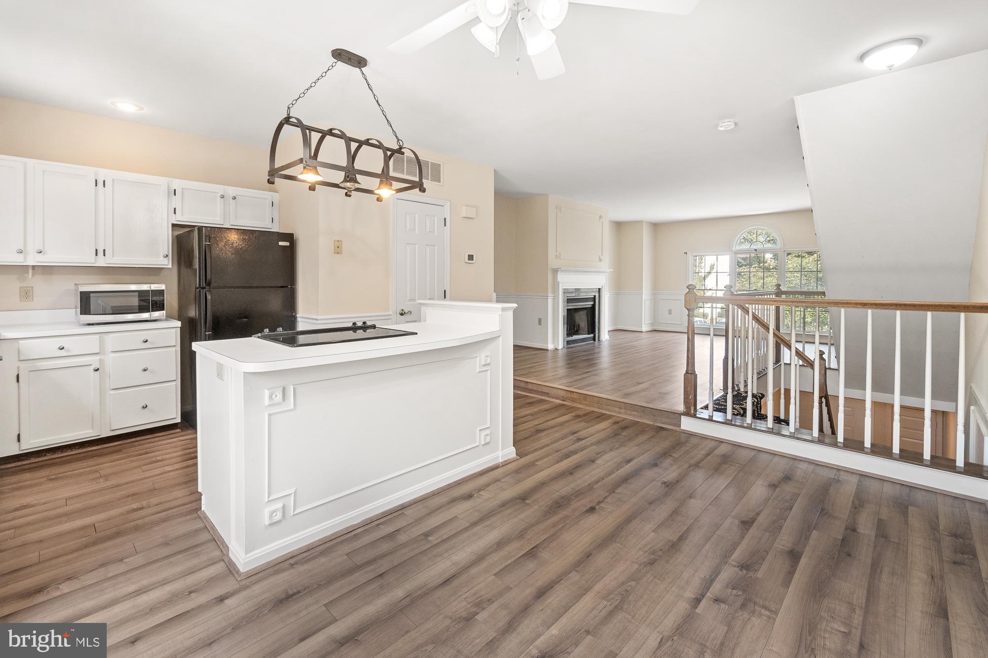 Open kitchen with white island and cooktop, wood-look floors, fireplace, and staircase railing.