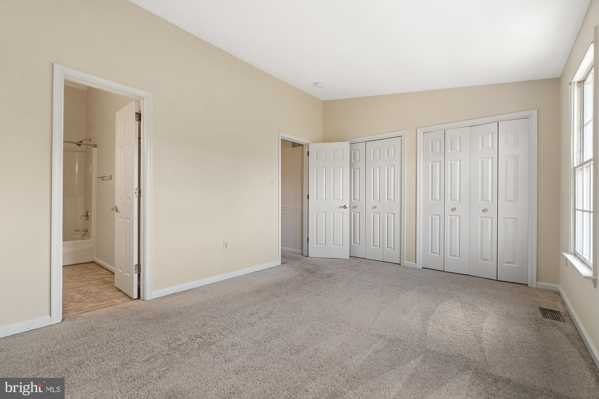Empty carpeted bedroom with white closets, a bright window, and an open doorway to the bathroom.