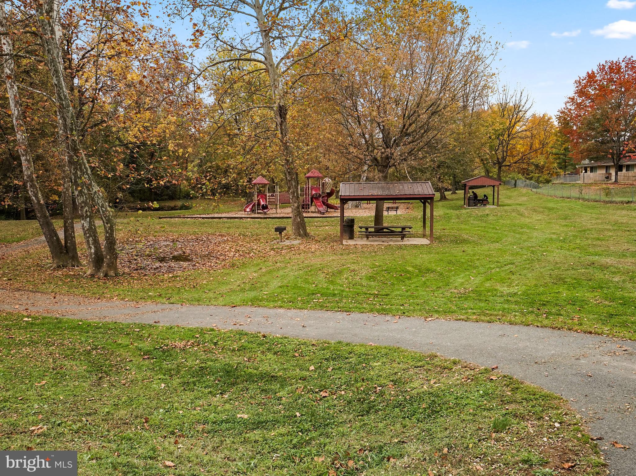 Neighborhood park with a red playground, picnic shelters, and a paved path beneath autumn trees.