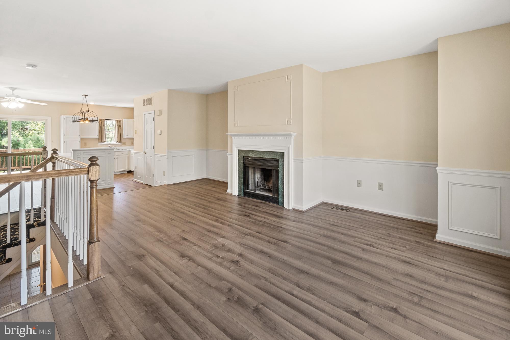 Open living area with a fireplace, wood-look floors, staircase railing, and kitchen beyond.