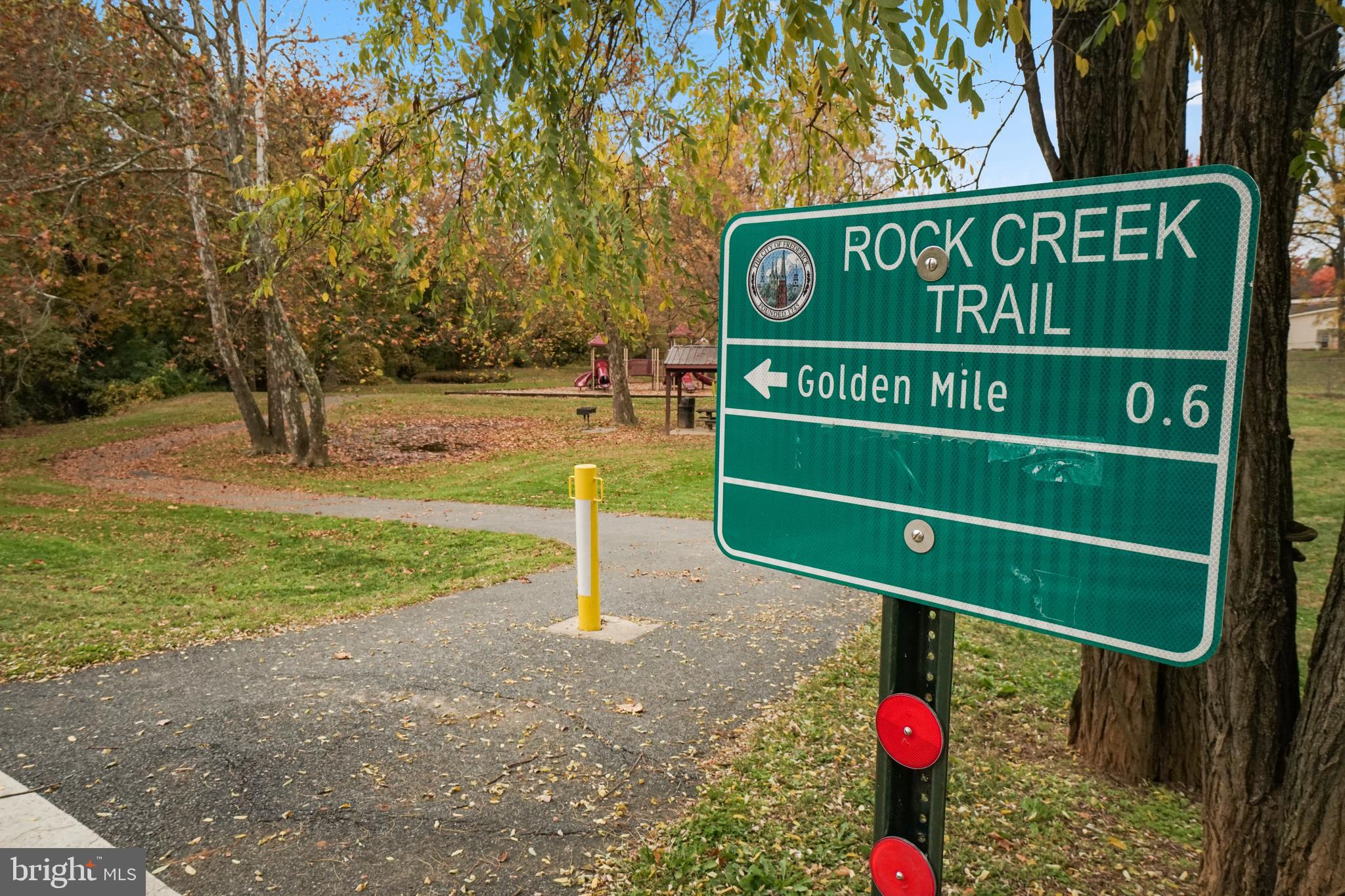 Rock Creek Trail sign beside a paved park path, with a playground and autumn trees.