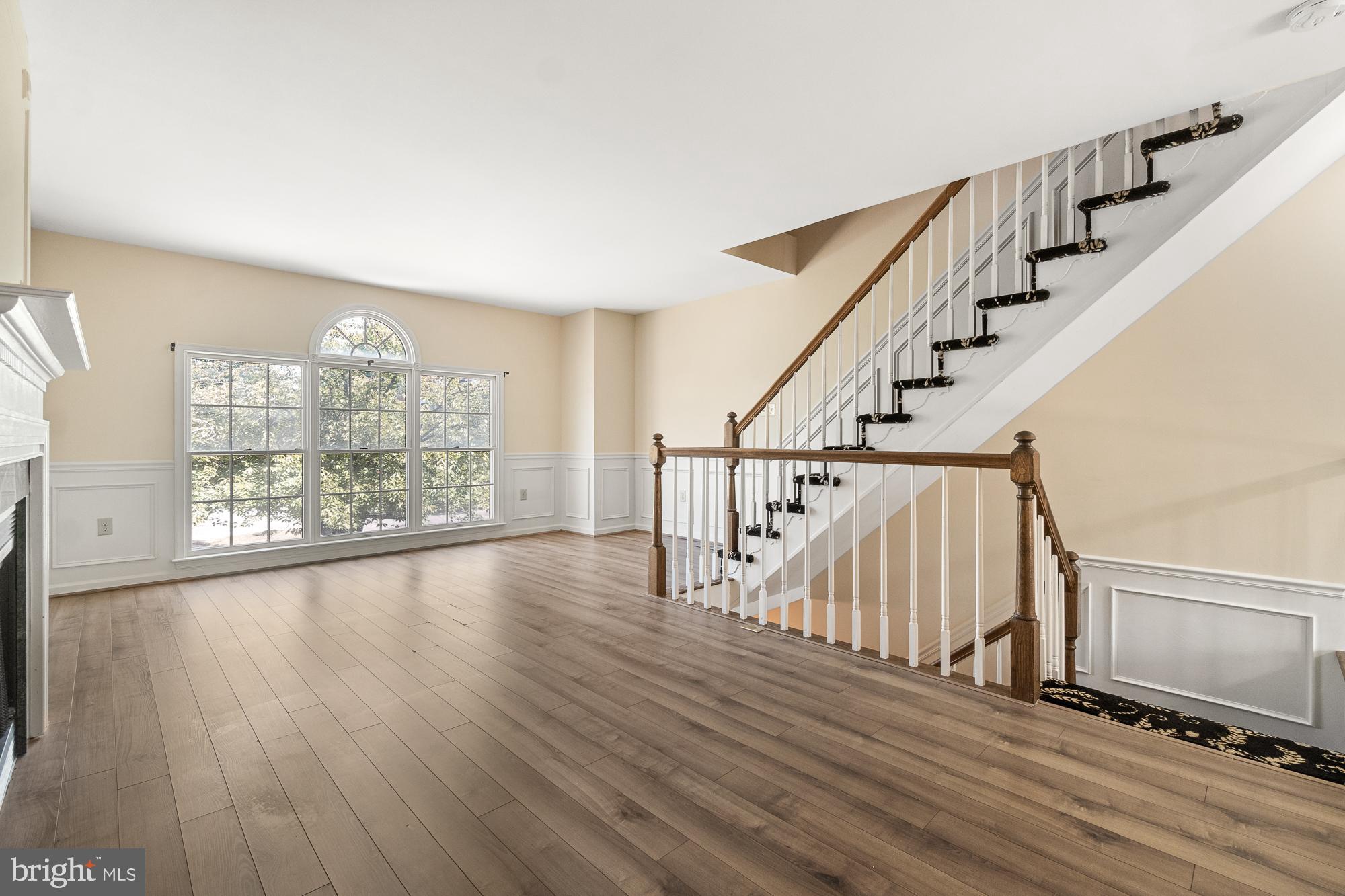 Empty living area with wide-plank flooring, a fireplace, large arched window, and prominent staircase.