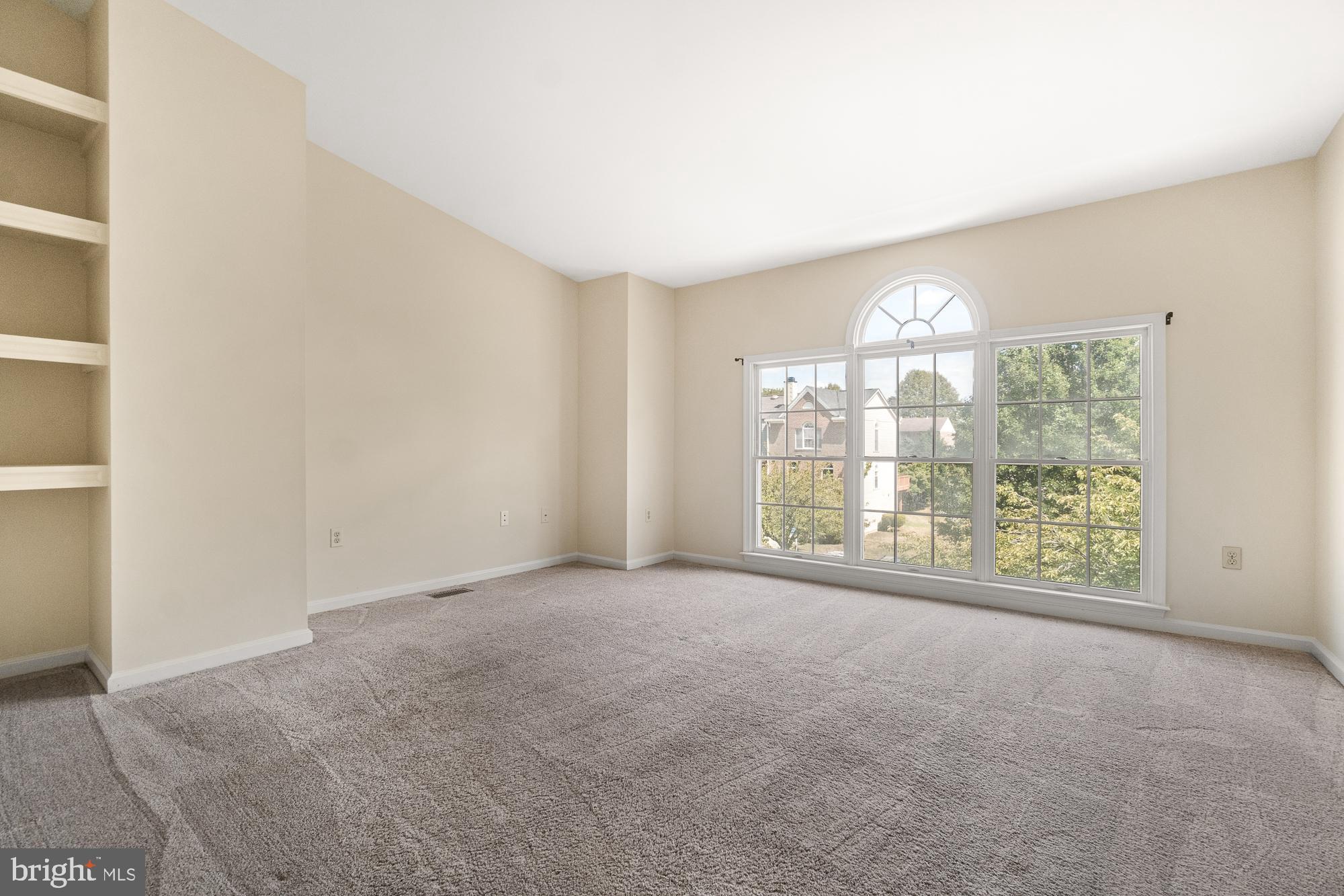Vacant living room with built-in shelves and large windows topped by an arched transom.