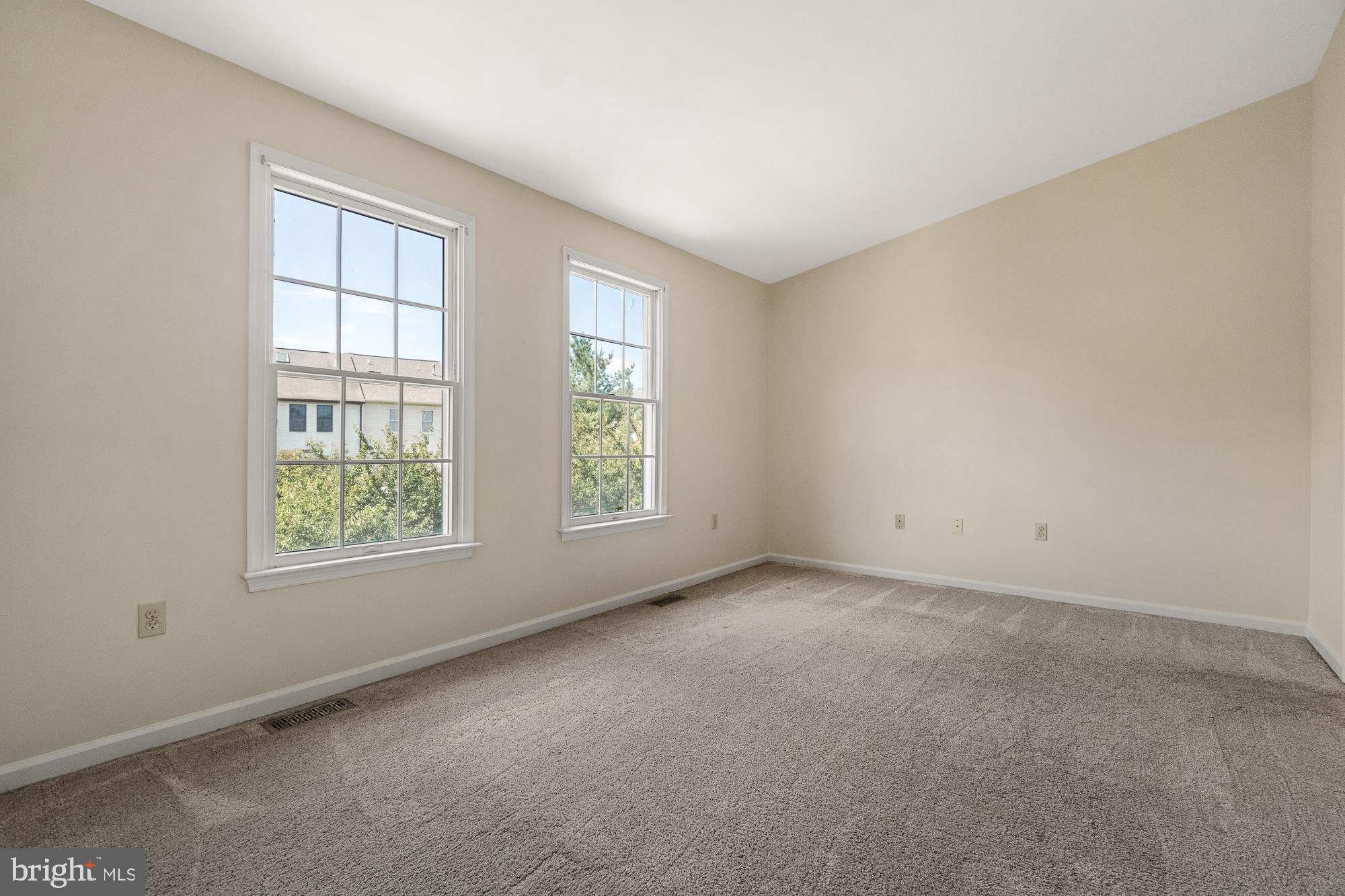Empty carpeted room with beige walls and two large windows.