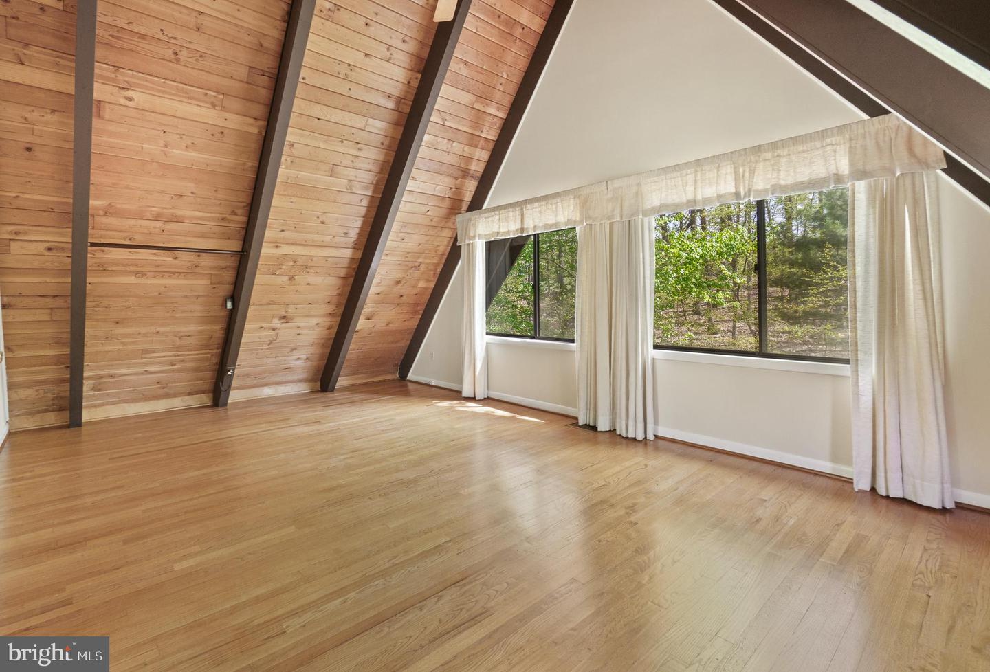 Vacant room with a sloped wood ceiling, exposed dark beams, hardwood floors, and wide curtained windows.
