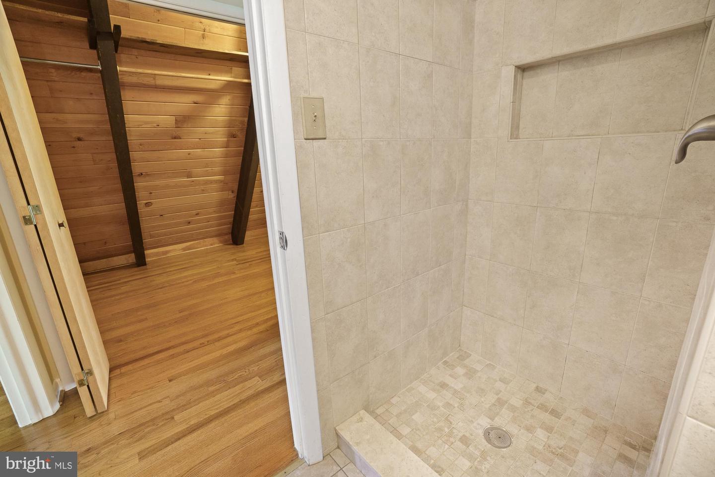Walk-in tiled shower with a recessed niche beside a wood-paneled room with hardwood floors.