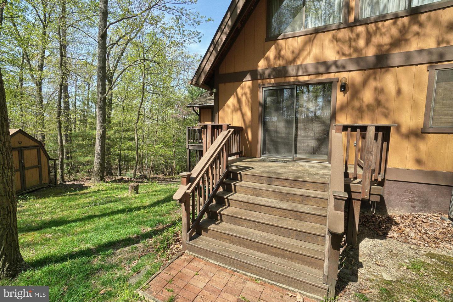 Brown-sided home with a raised wooden deck, brick patio, and wooded yard.