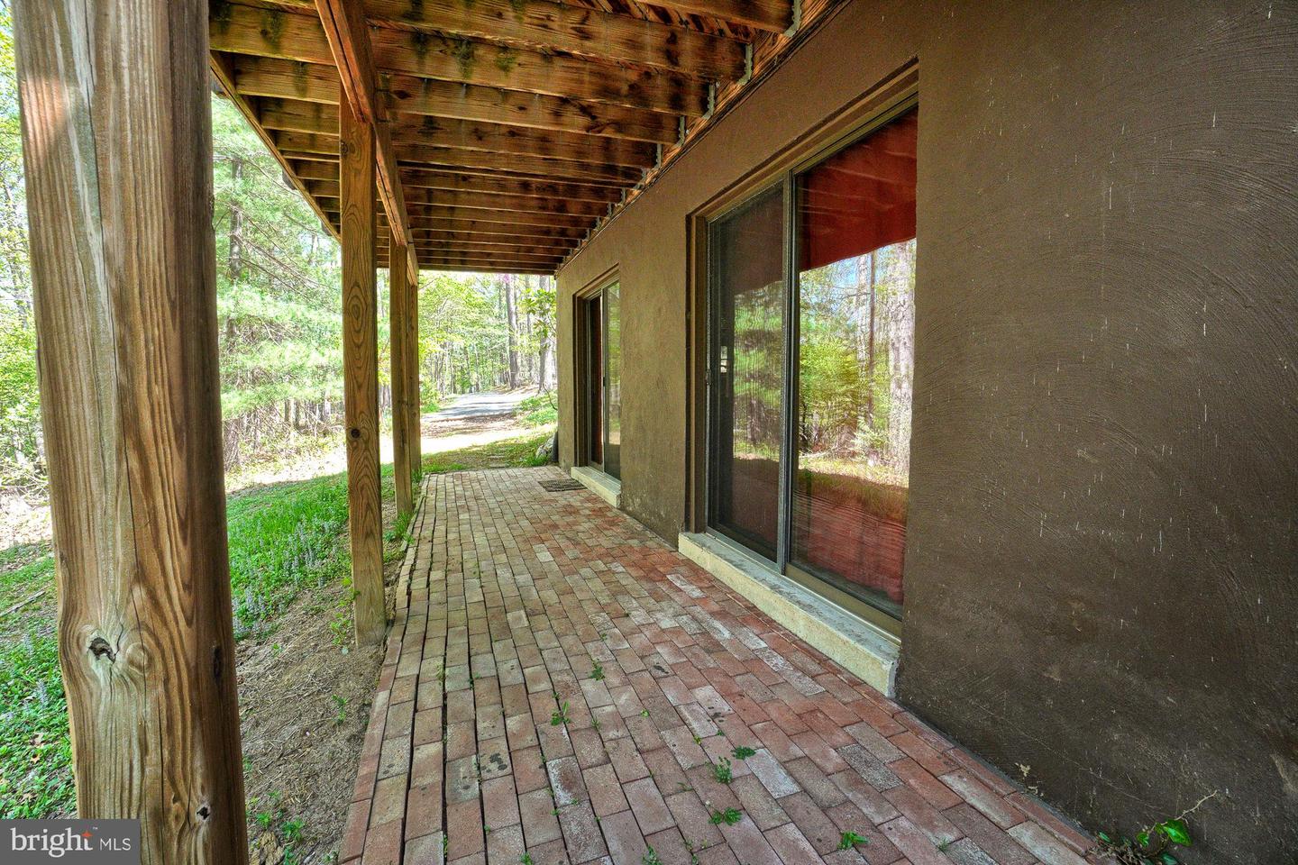 Covered brick patio with timber posts and sliding glass doors beside a wooded driveway.