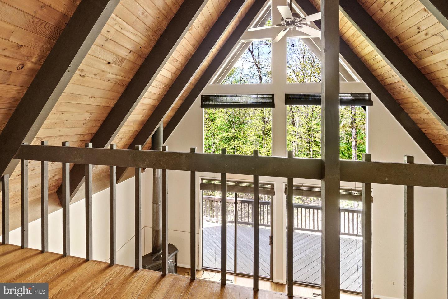 Vaulted wood-paneled loft with dark beams, a ceiling fan, and large windows overlooking trees and a deck.