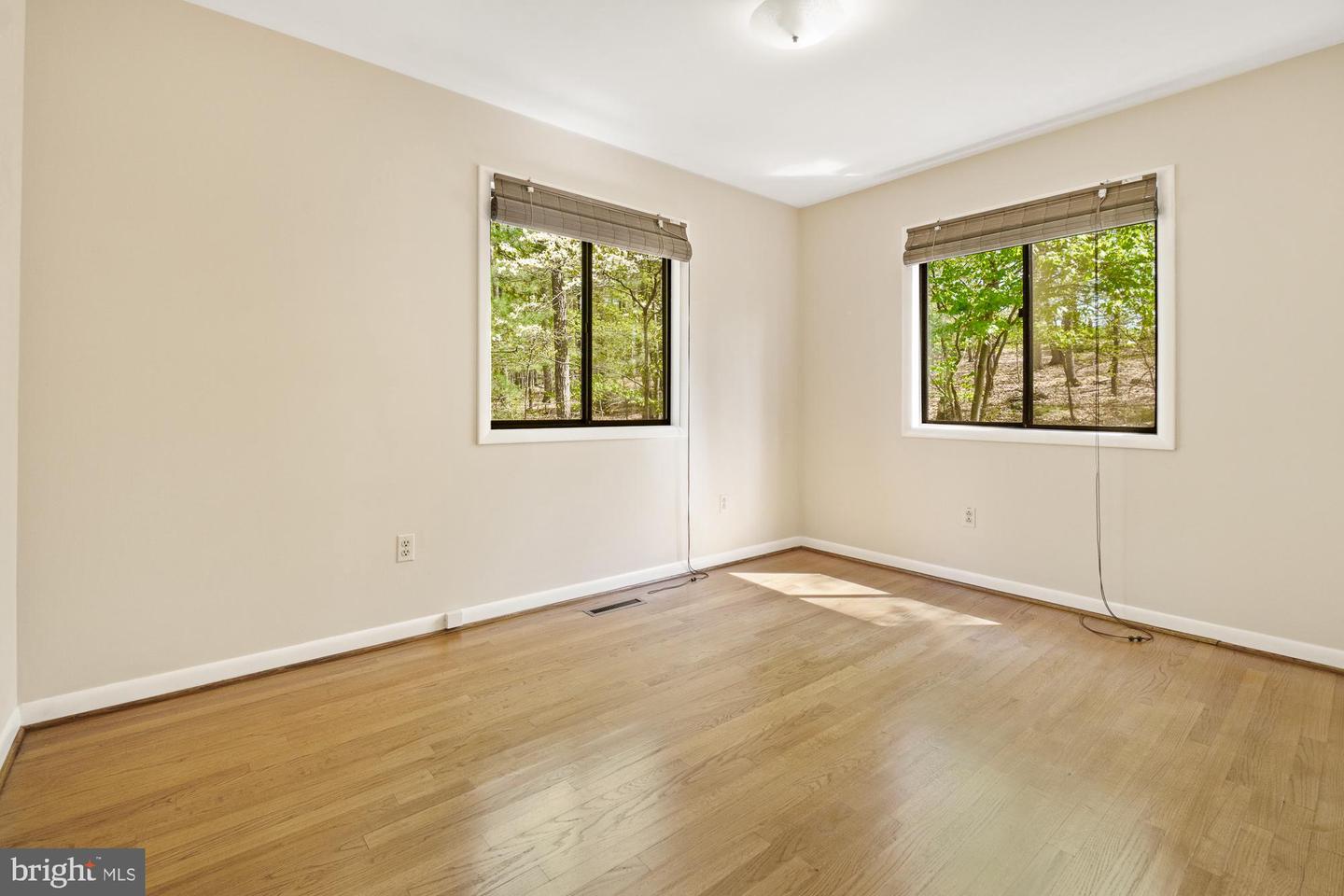 Empty room with hardwood floors and two windows overlooking leafy trees.