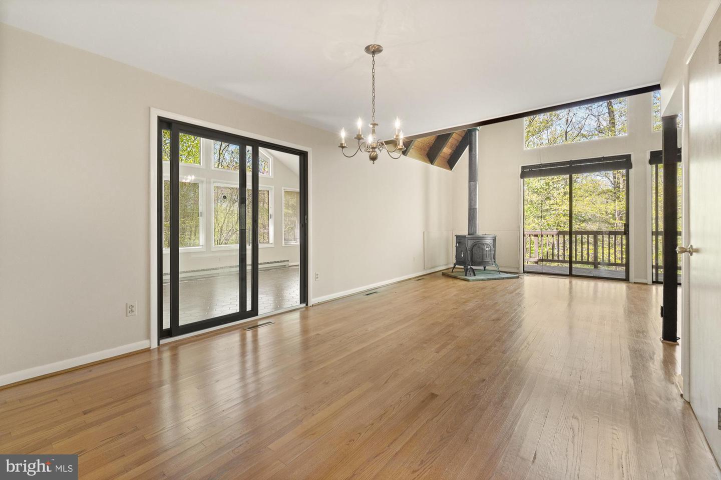 Empty living room with hardwood floors, a freestanding wood stove, and large windows overlooking a wooded deck.