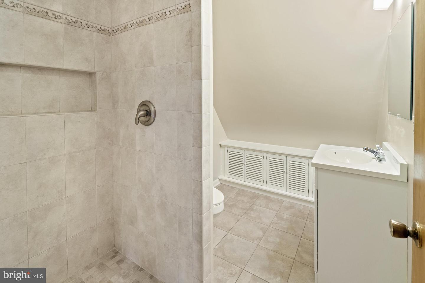 Bathroom with a beige tiled shower, recessed niche, white vanity, and louvered built-in cabinetry.