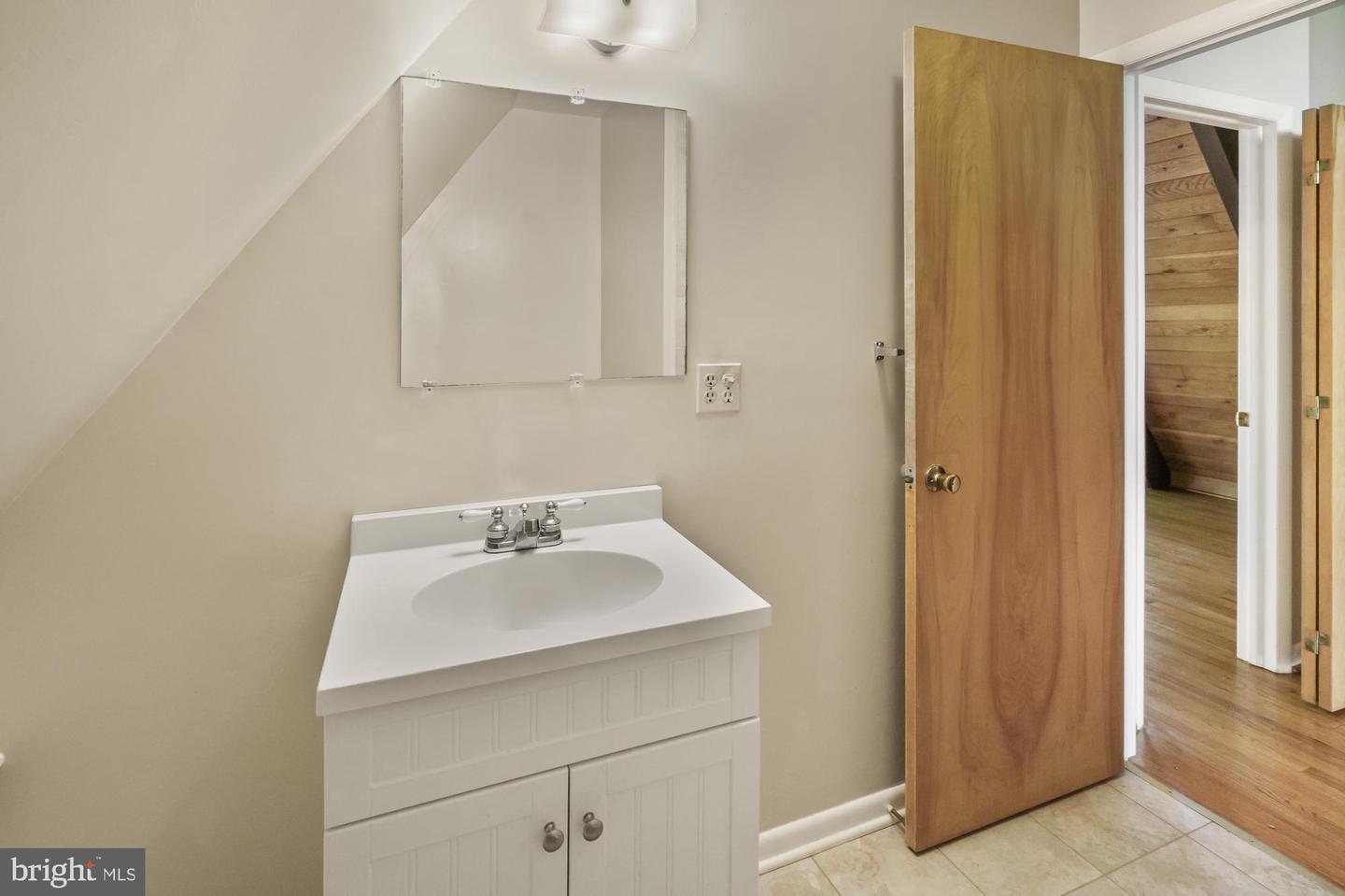 Bathroom vanity and mirror beneath a sloped ceiling, with an open door to a wood-floored room.