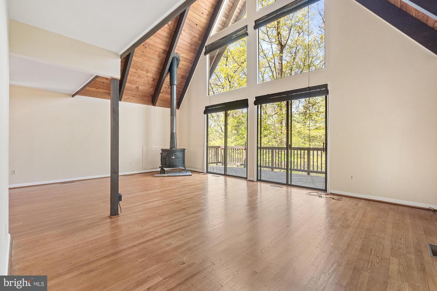 Vacant living room with vaulted wood ceiling, hardwood floors, wood stove, and windows overlooking a deck.