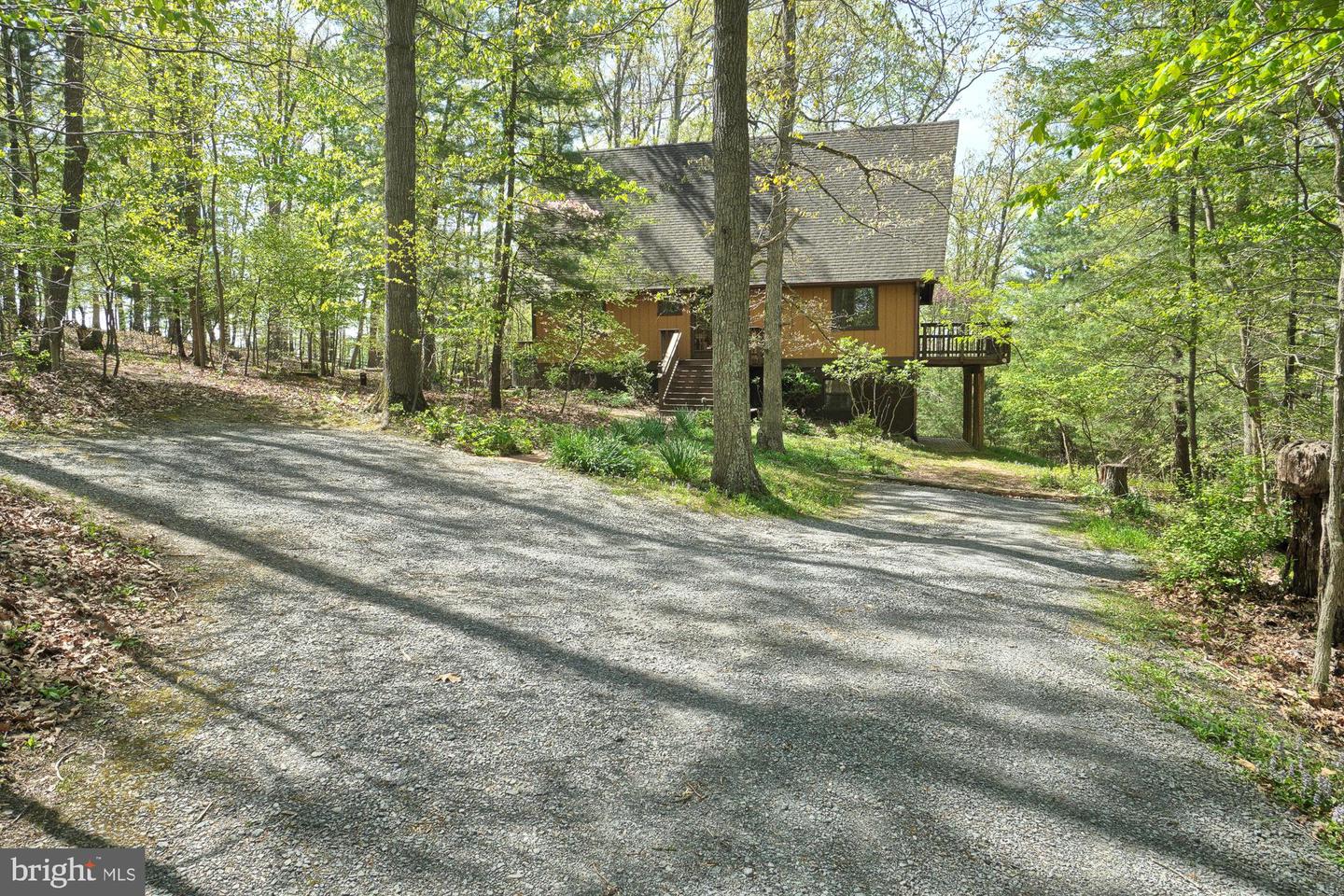 Wooded raised home with a steep roof, side deck, and broad gravel driveway.