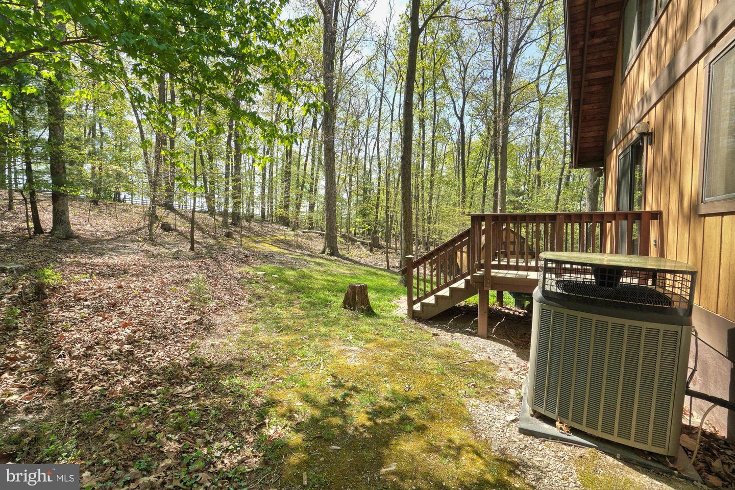 Wooded side yard beside a home, with a wooden deck, steps, and outdoor HVAC unit.