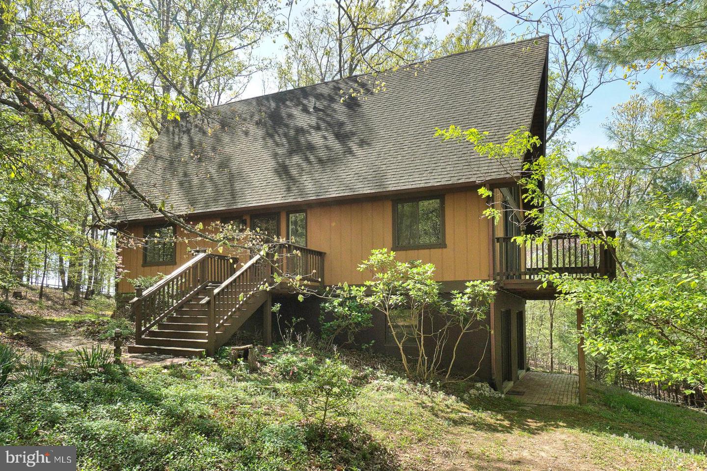 Raised brown house with a steep gabled roof, wooden stairs, and deck in a wooded setting.