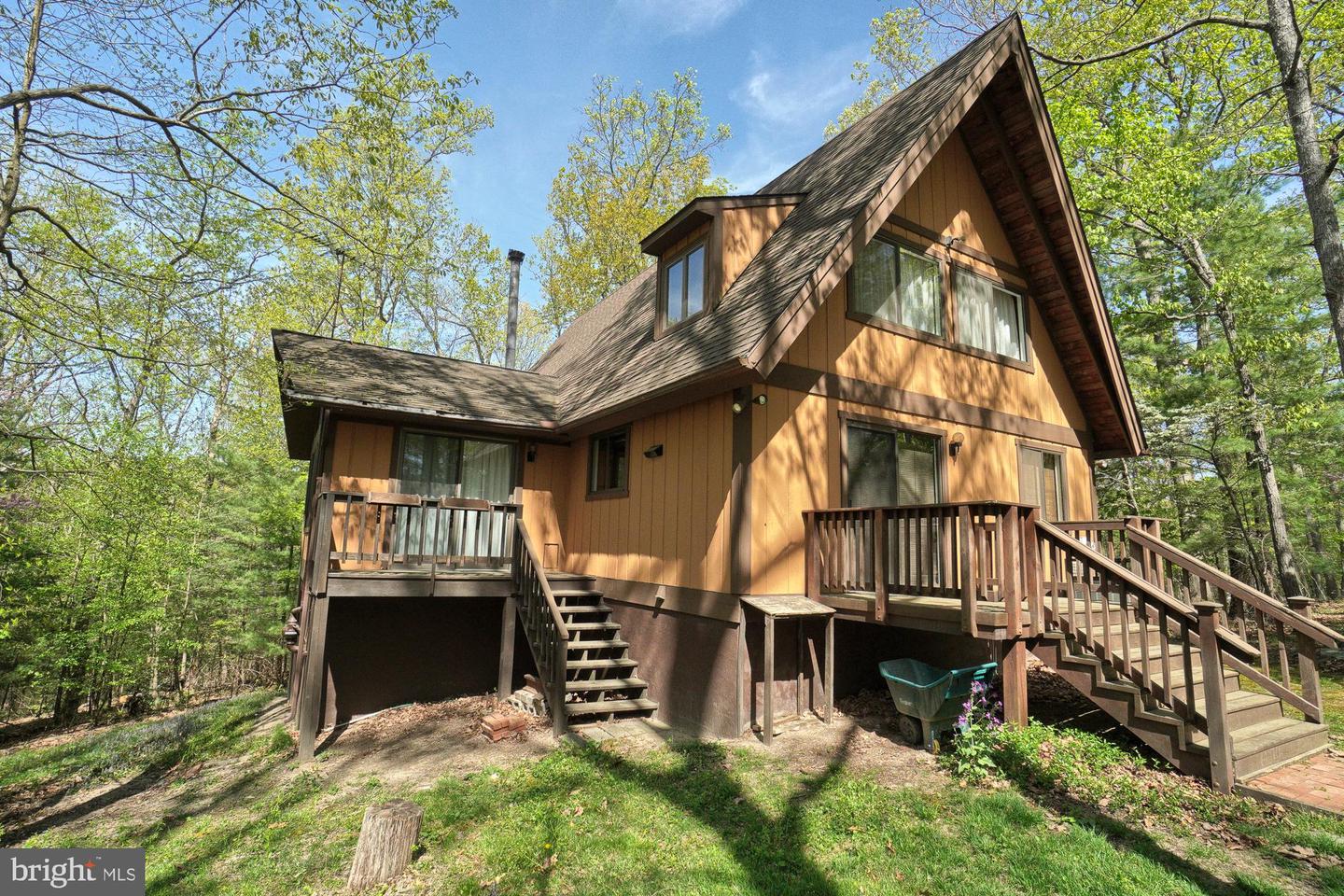 Wood-sided A-frame house with elevated decks and stairs in a leafy wooded setting.