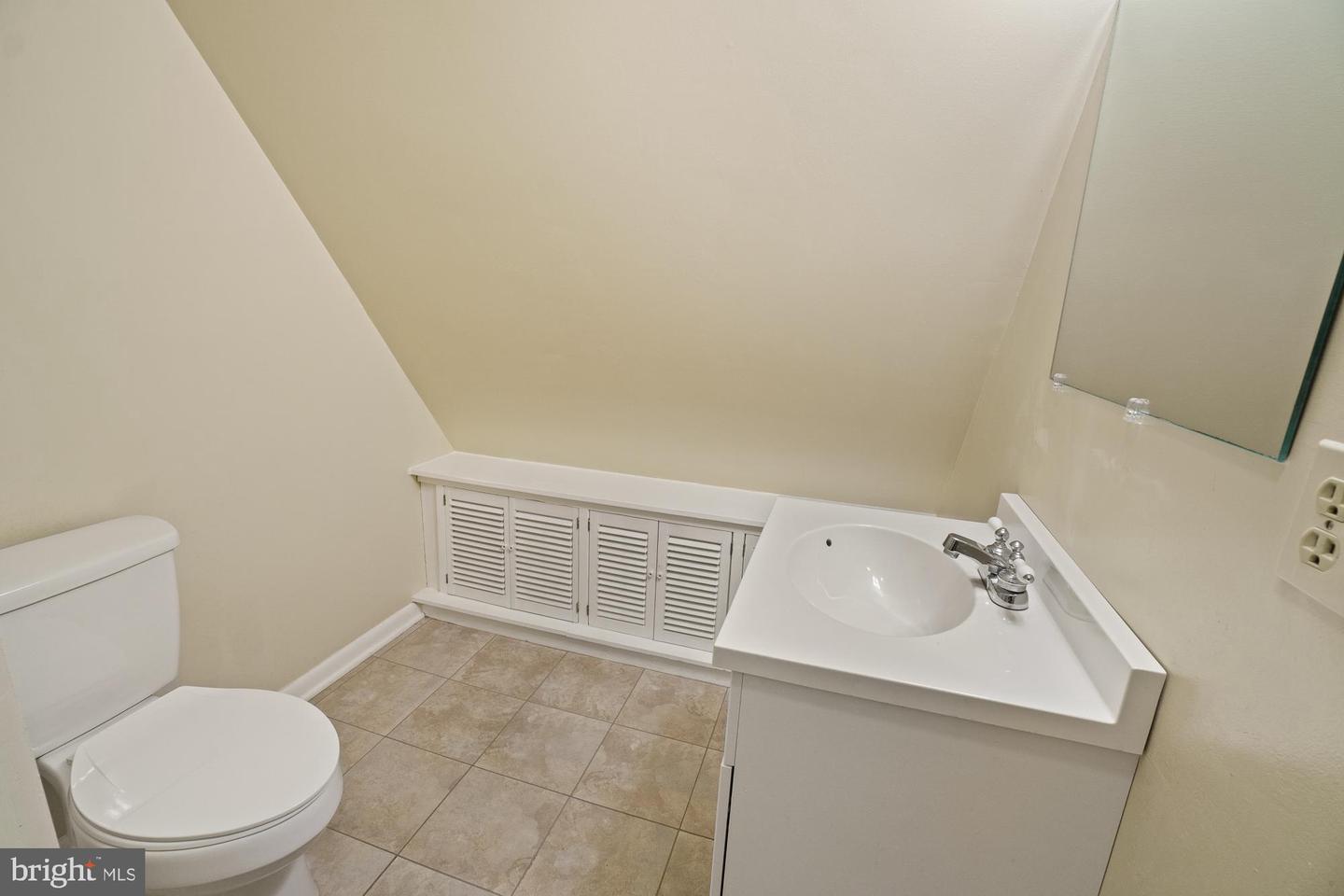 Bathroom with a white vanity and toilet, beige tile floor, and built-in louvered cabinets beneath a sloped ceiling.