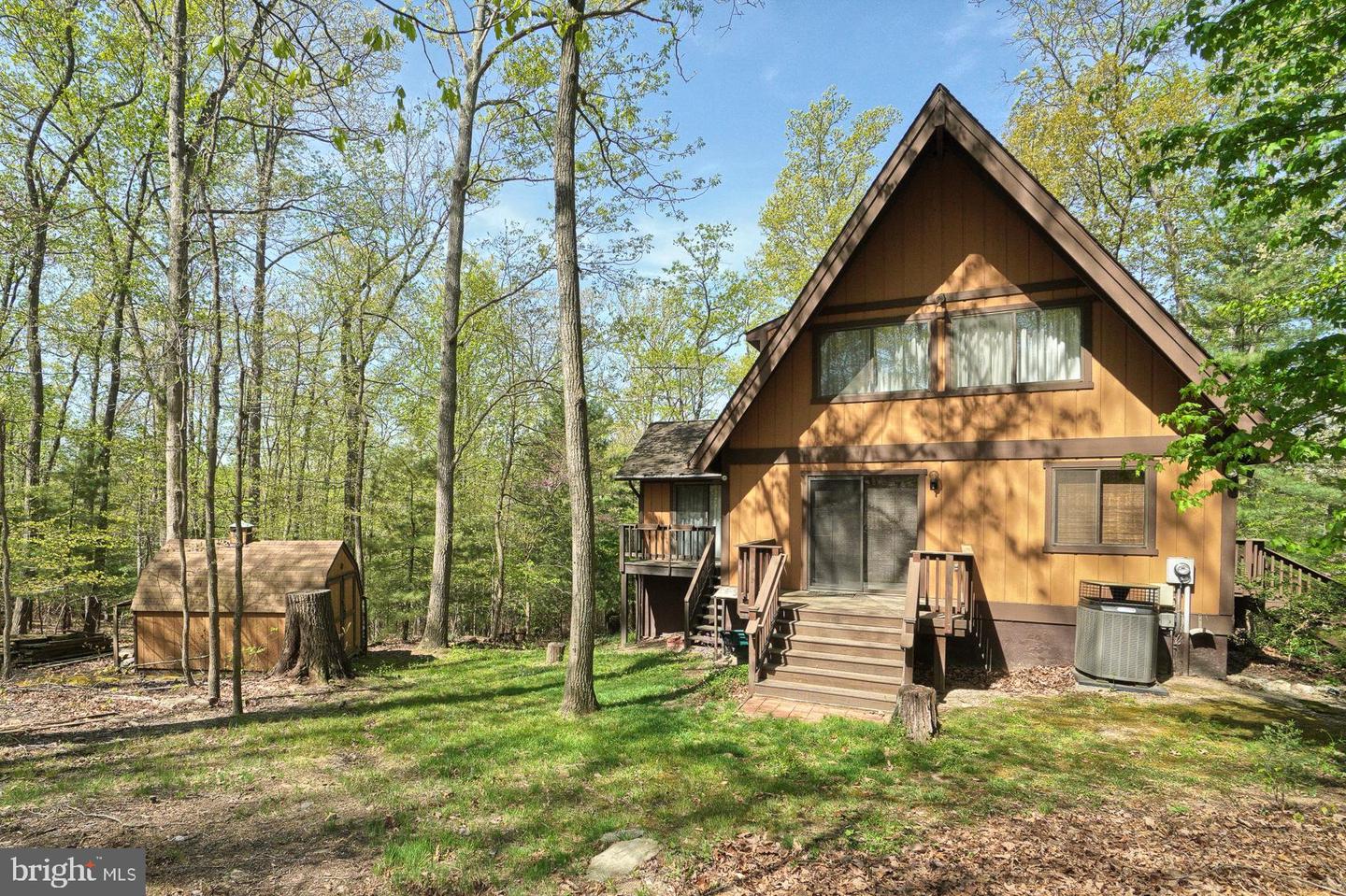 Brown A-frame house with a raised rear deck and stairs in a wooded yard beside a small shed.