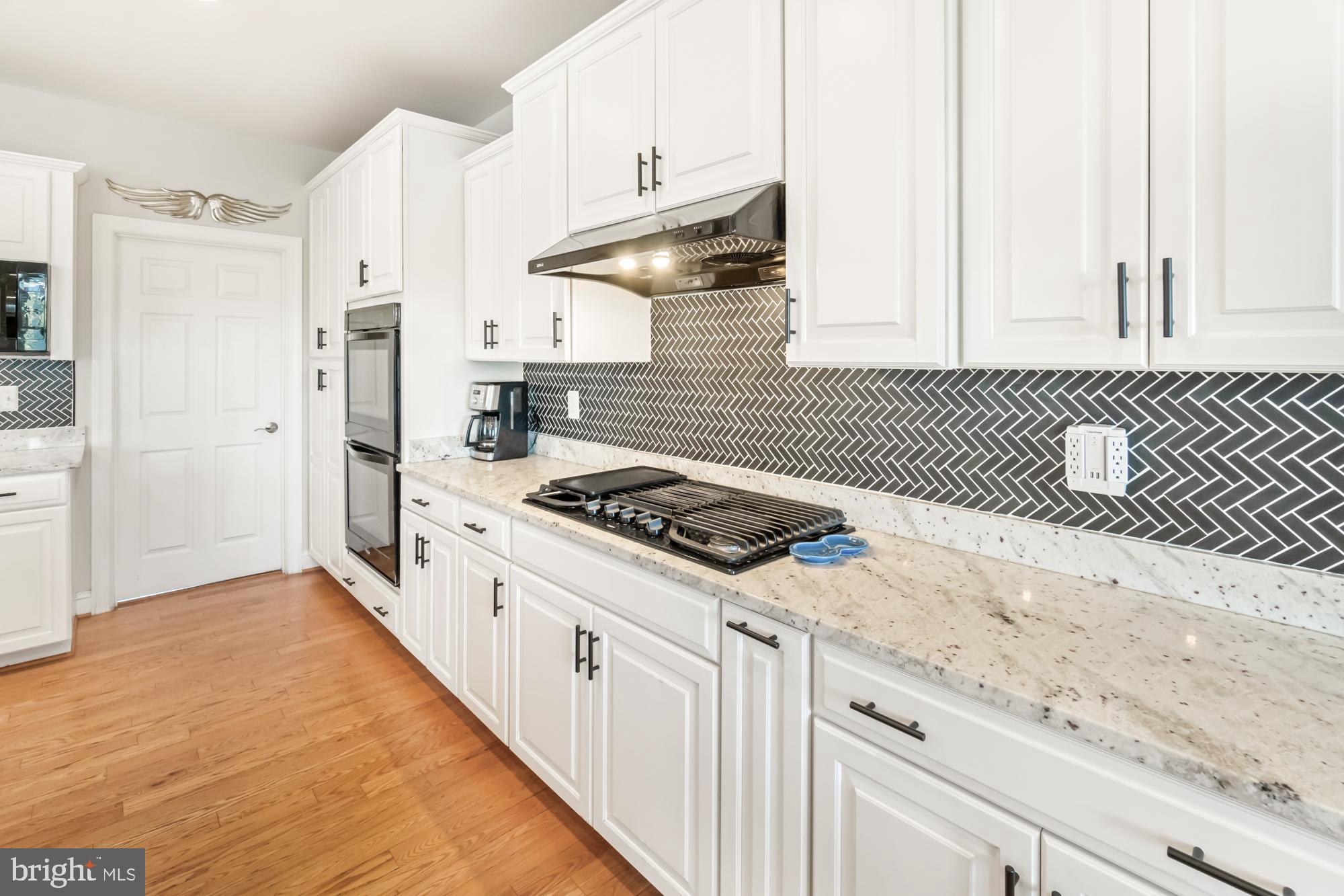 White kitchen with dark herringbone backsplash, stone counters, gas cooktop, and hardwood floors.