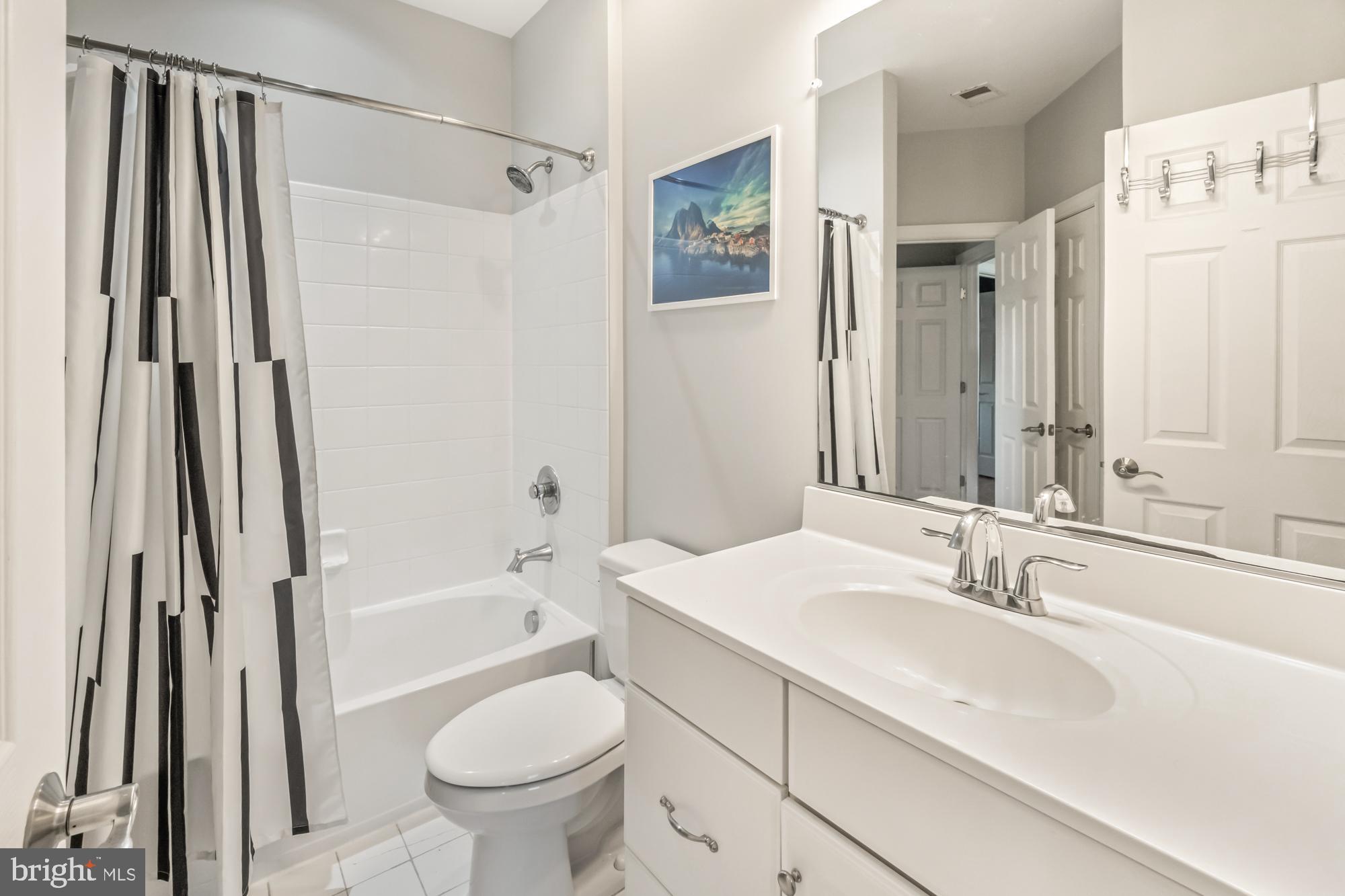 Bathroom with a white tub, geometric shower curtain, toilet, and white vanity with a large mirror.