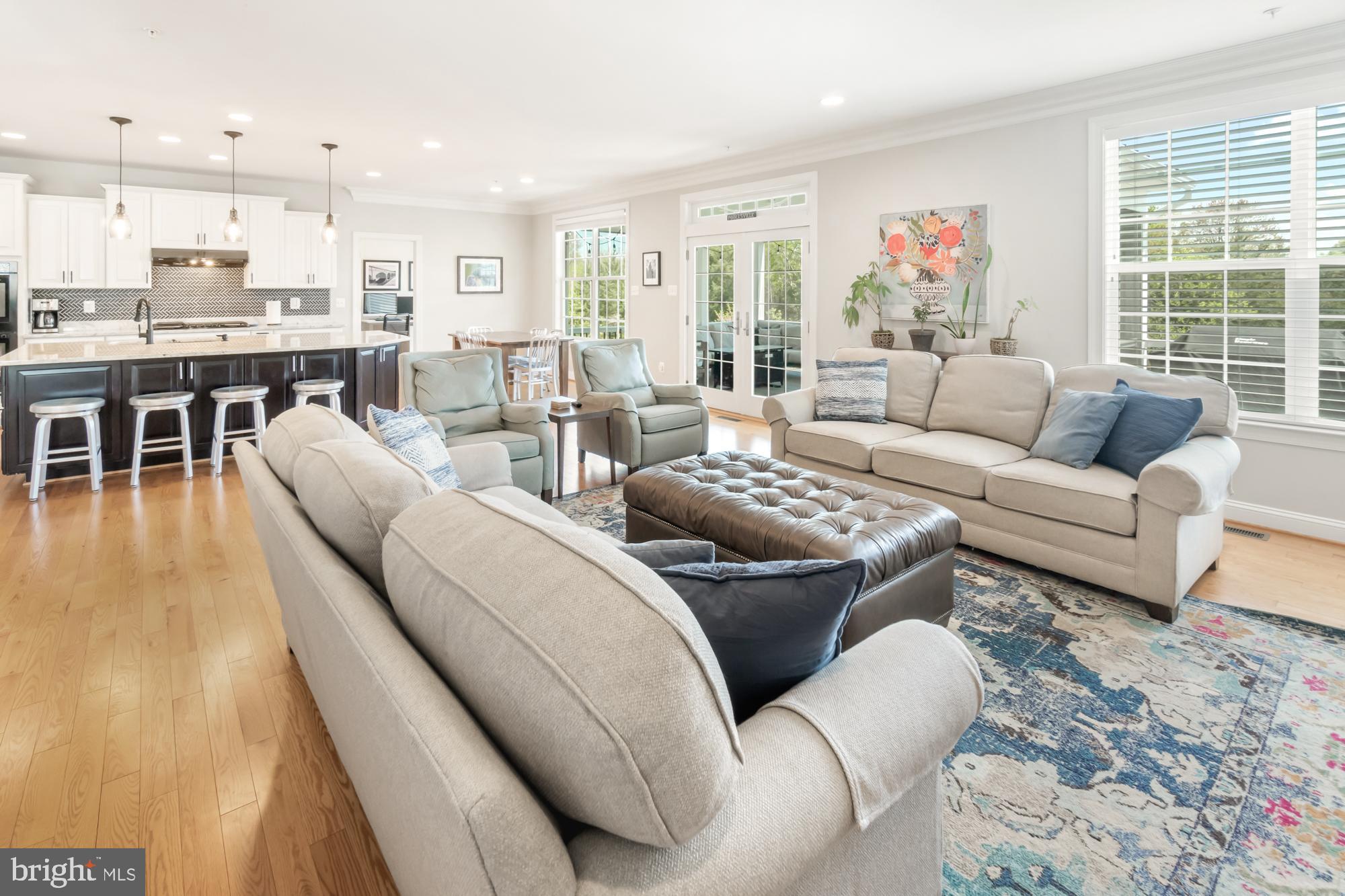 Open-plan living room and kitchen with a large sectional, leather ottoman, and white kitchen cabinets.