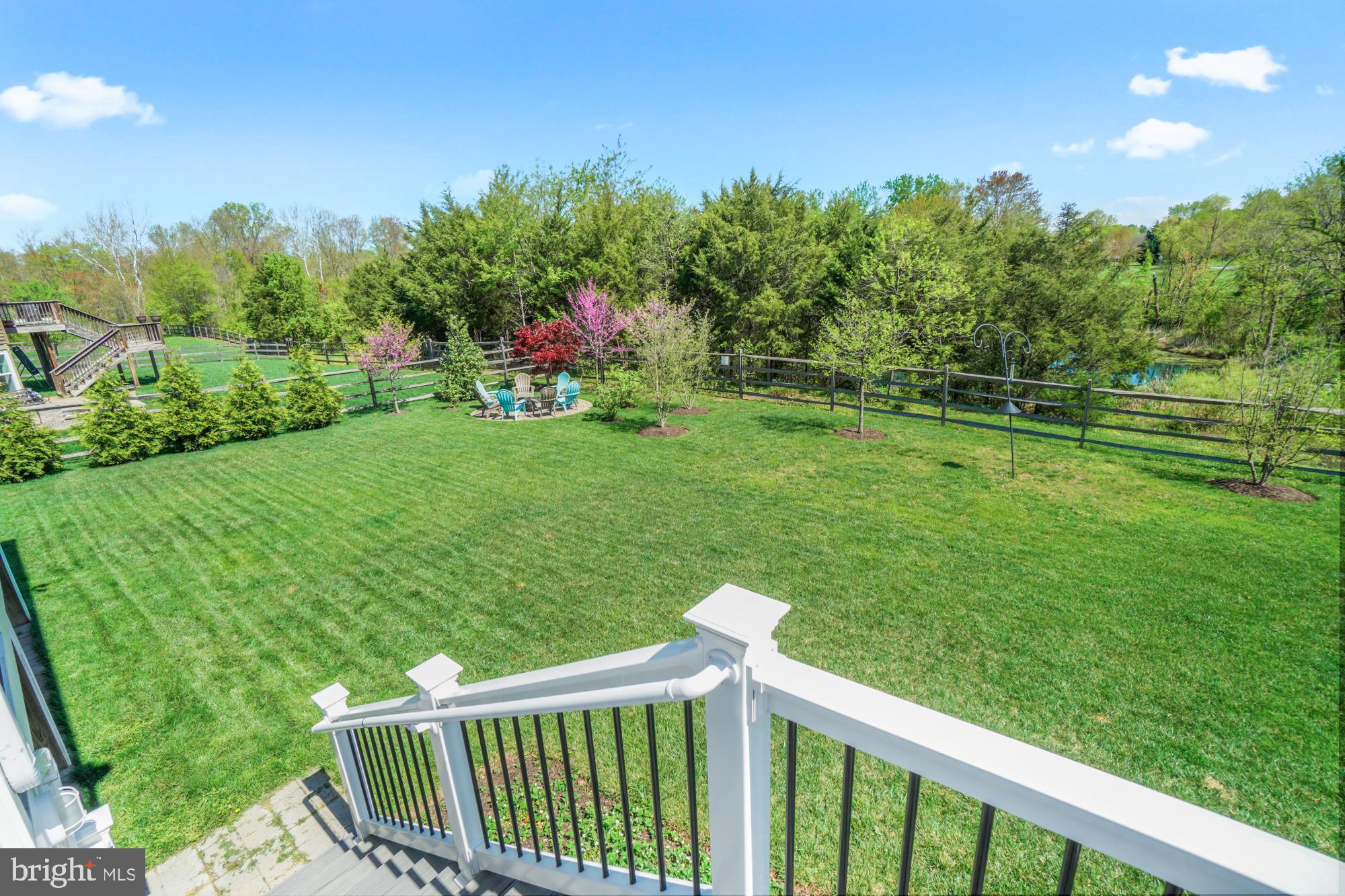 Fenced backyard with a broad lawn, flowering trees, outdoor seating, and a pond beyond the fence.