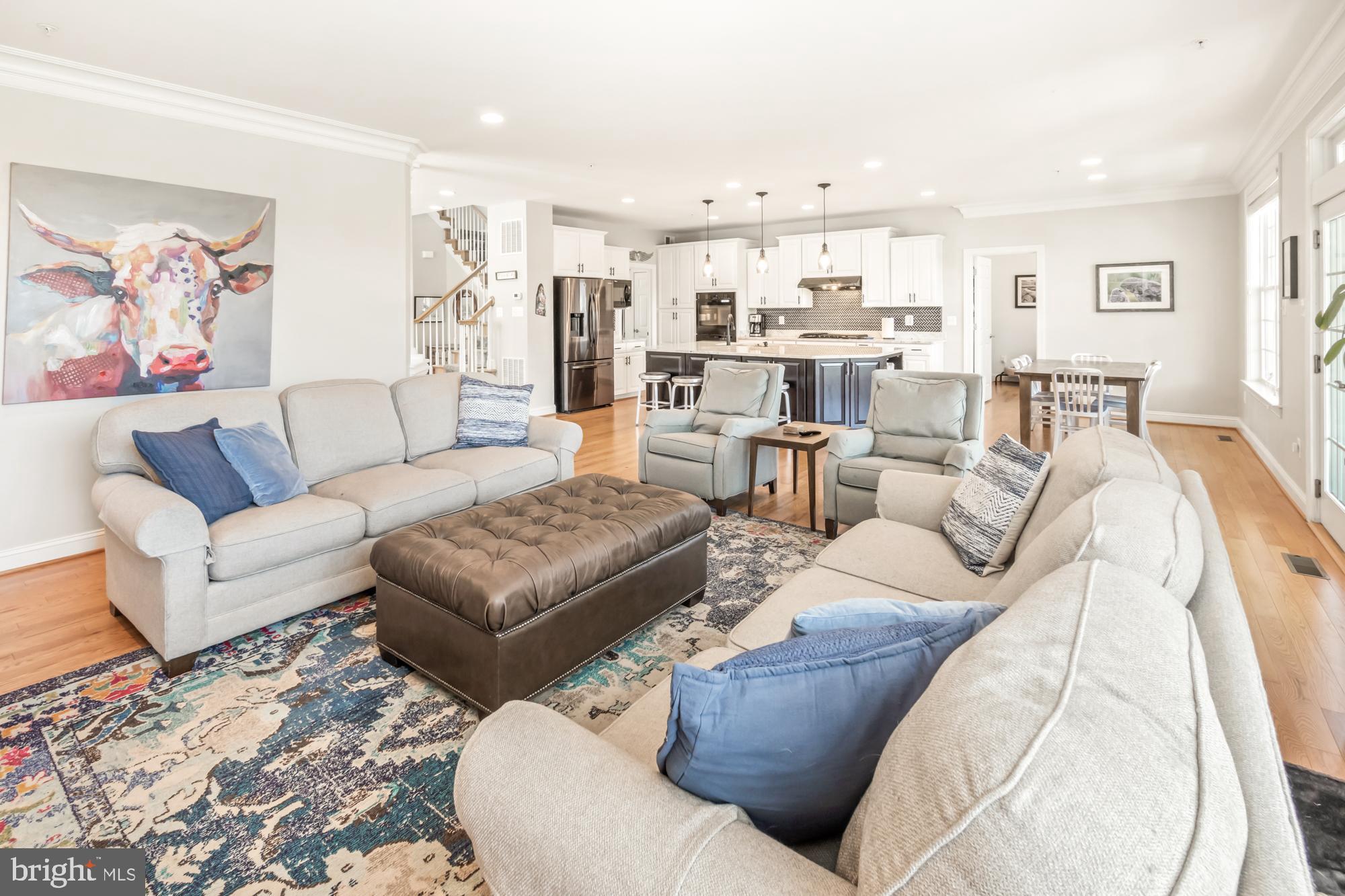 Open-plan living room with pale sofas and a leather ottoman, adjoining a white kitchen and dining area.
