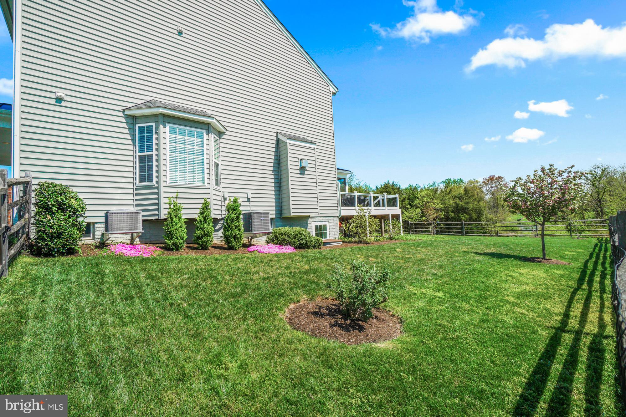 House beside a broad fenced lawn, with a raised deck, bay window, and flowering trees.