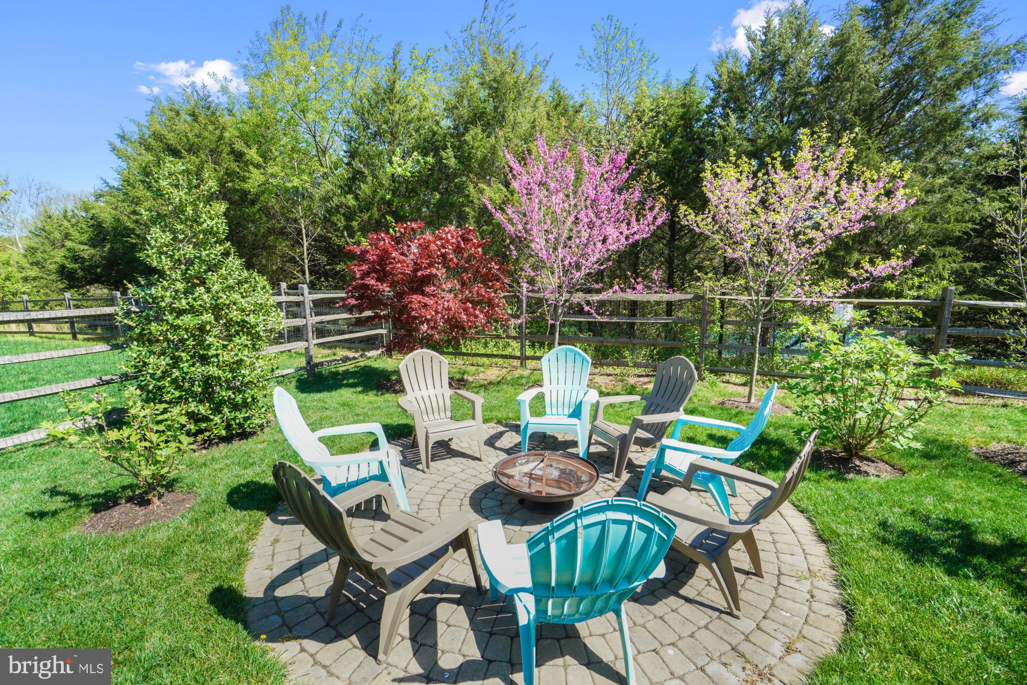 Stone patio with Adirondack chairs around a fire pit, beside a fenced lawn and flowering trees.