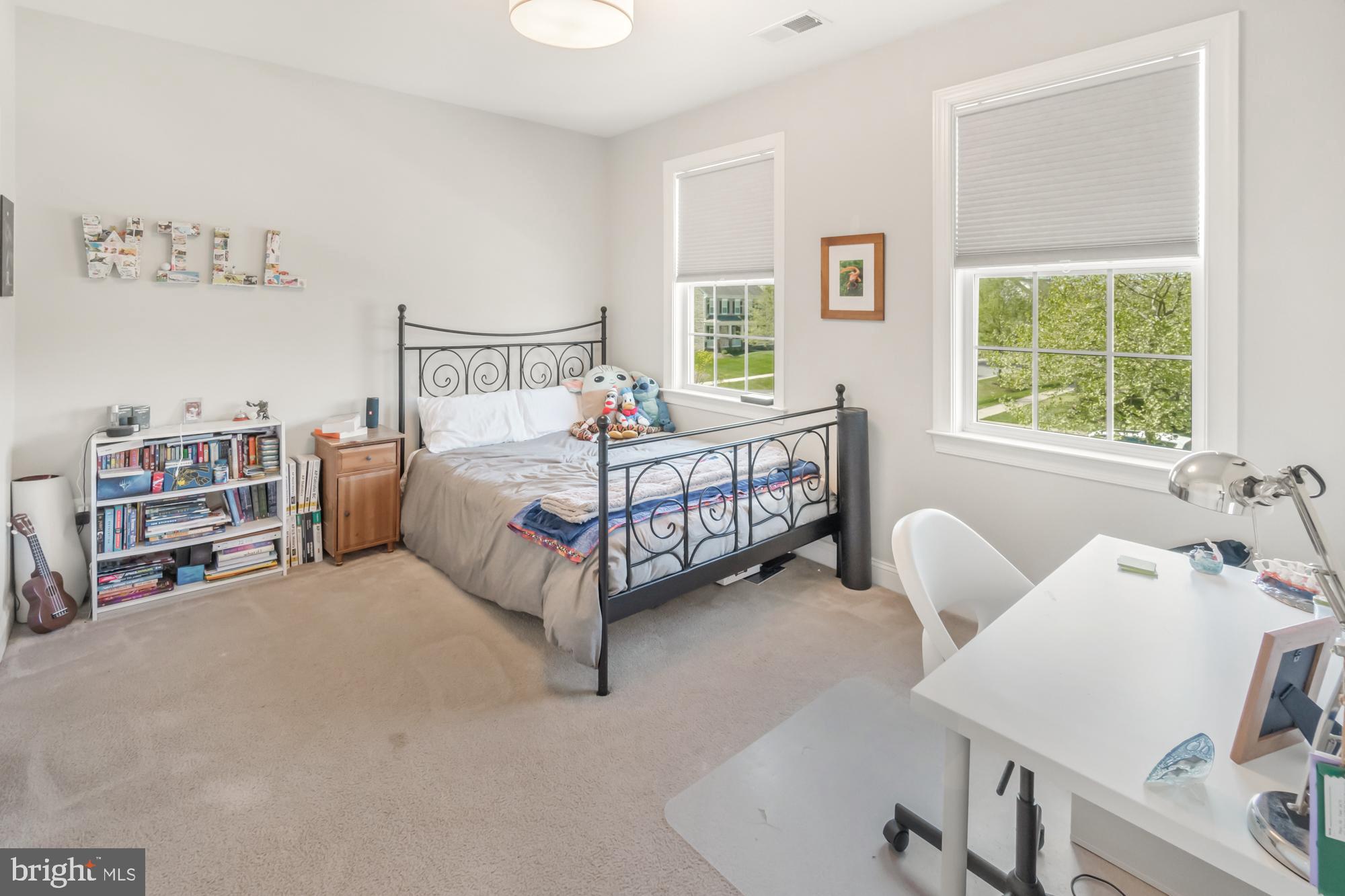 Bedroom with a black metal bed, plush toys, two large windows, and a white desk.
