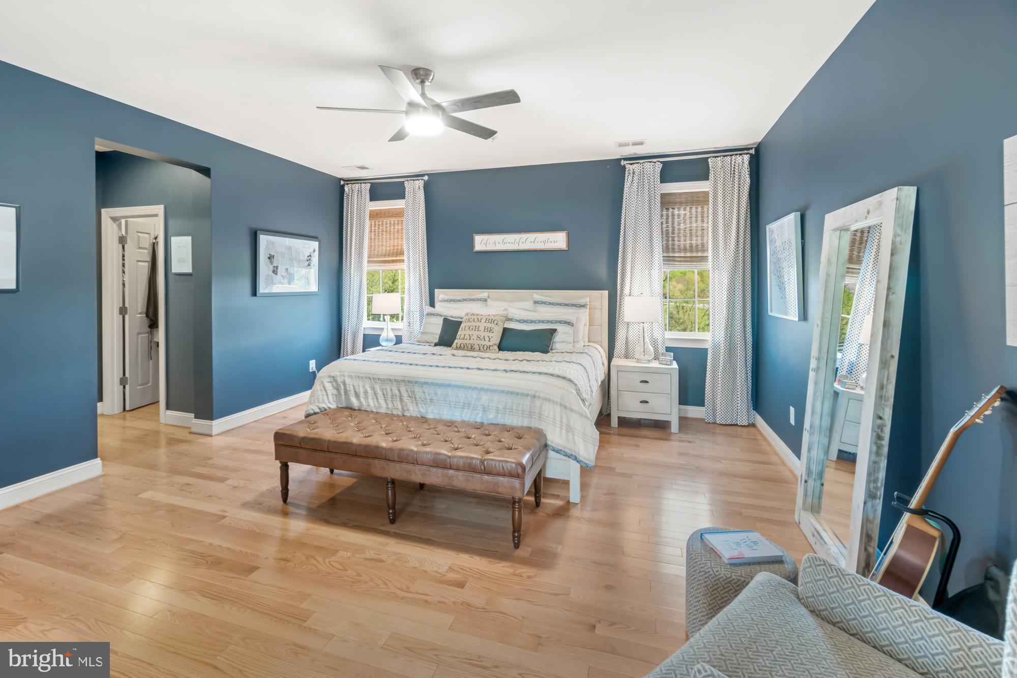 Furnished bedroom with blue walls, a white bed, leather bench, and leaning full-length mirror.