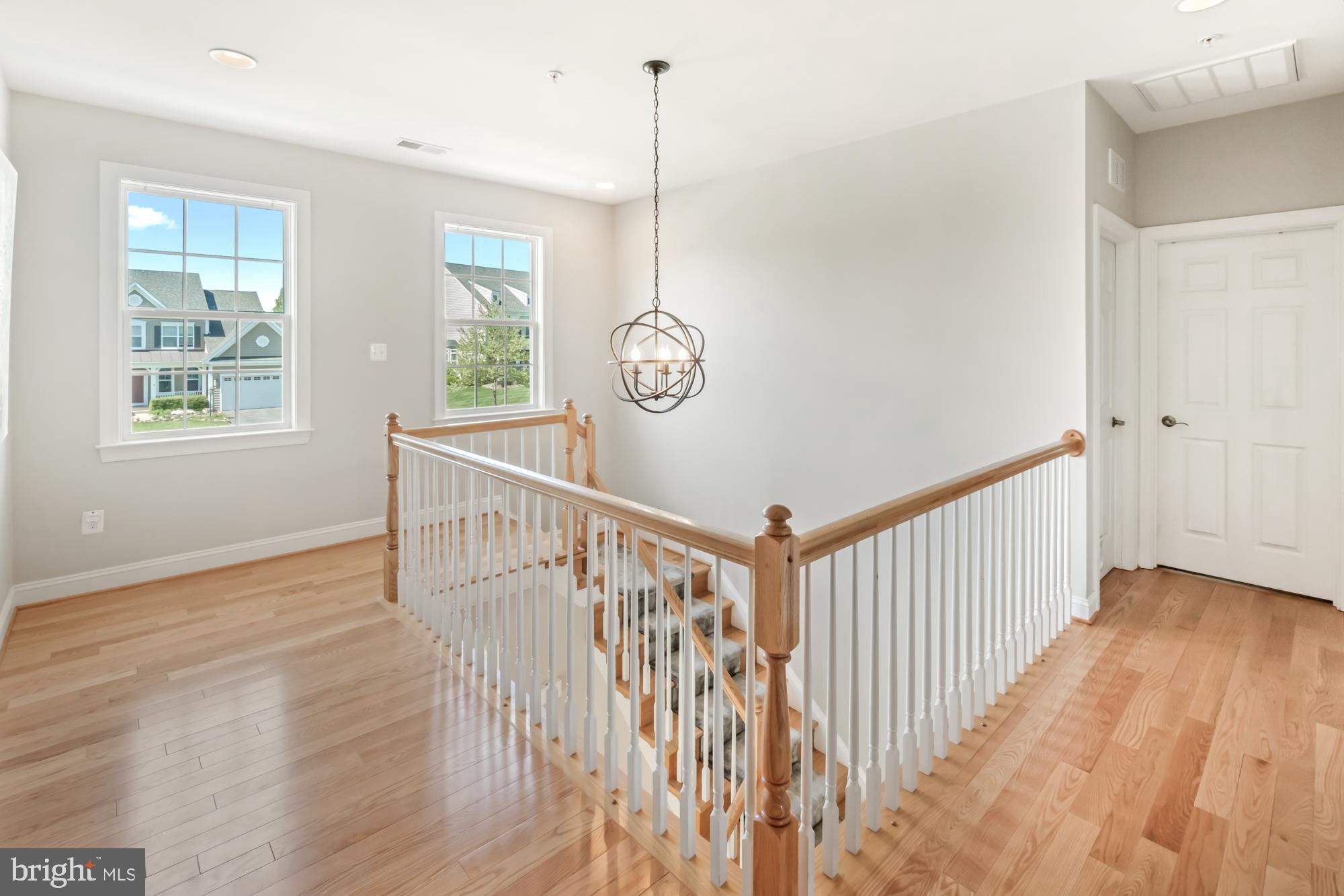 Upstairs landing with hardwood floors, a wood-and-white stair railing, two windows, and a hanging chandelier.