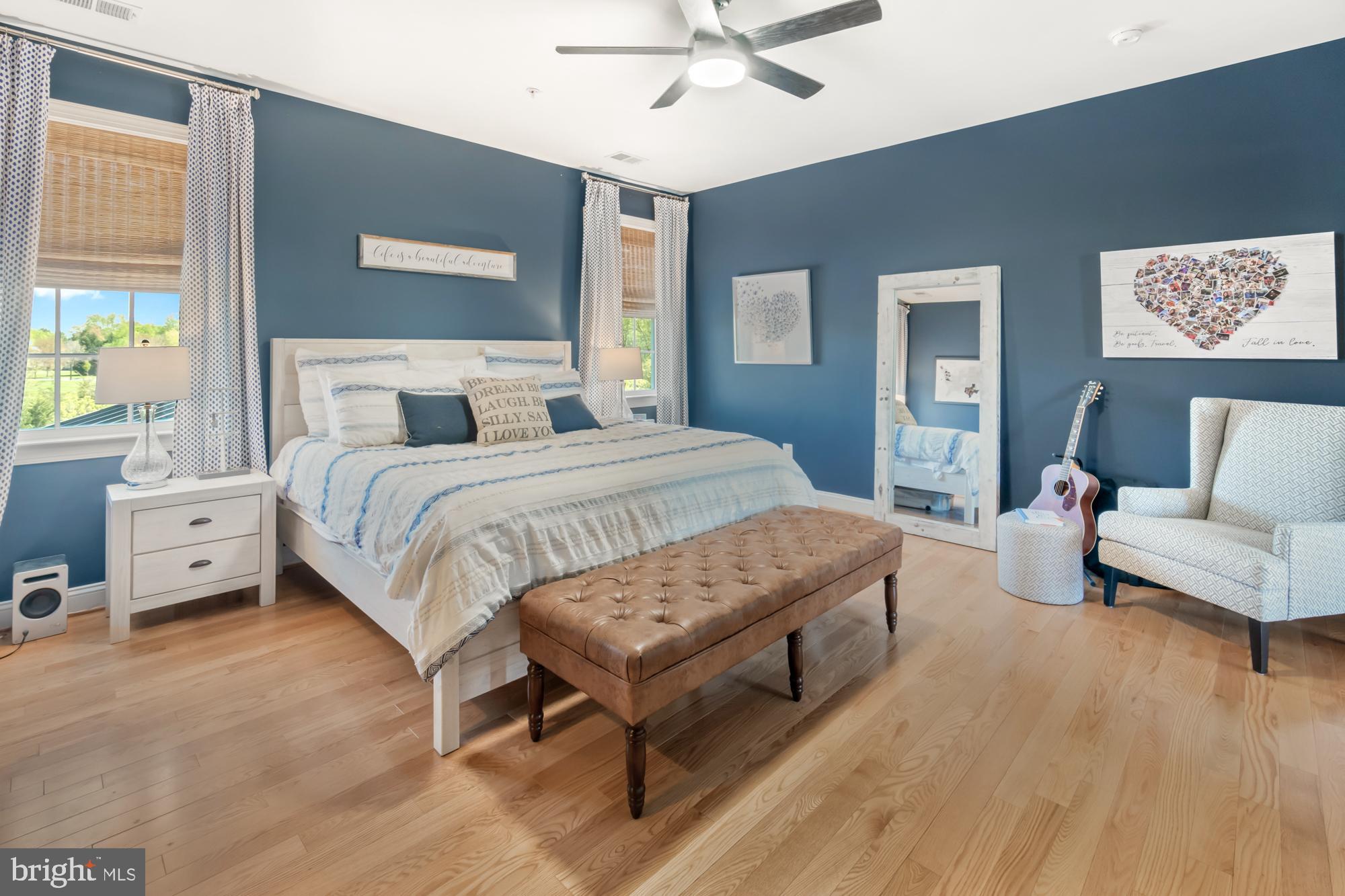 Blue bedroom with a white bed, tufted brown bench, hardwood floors, and guitar beside an armchair.