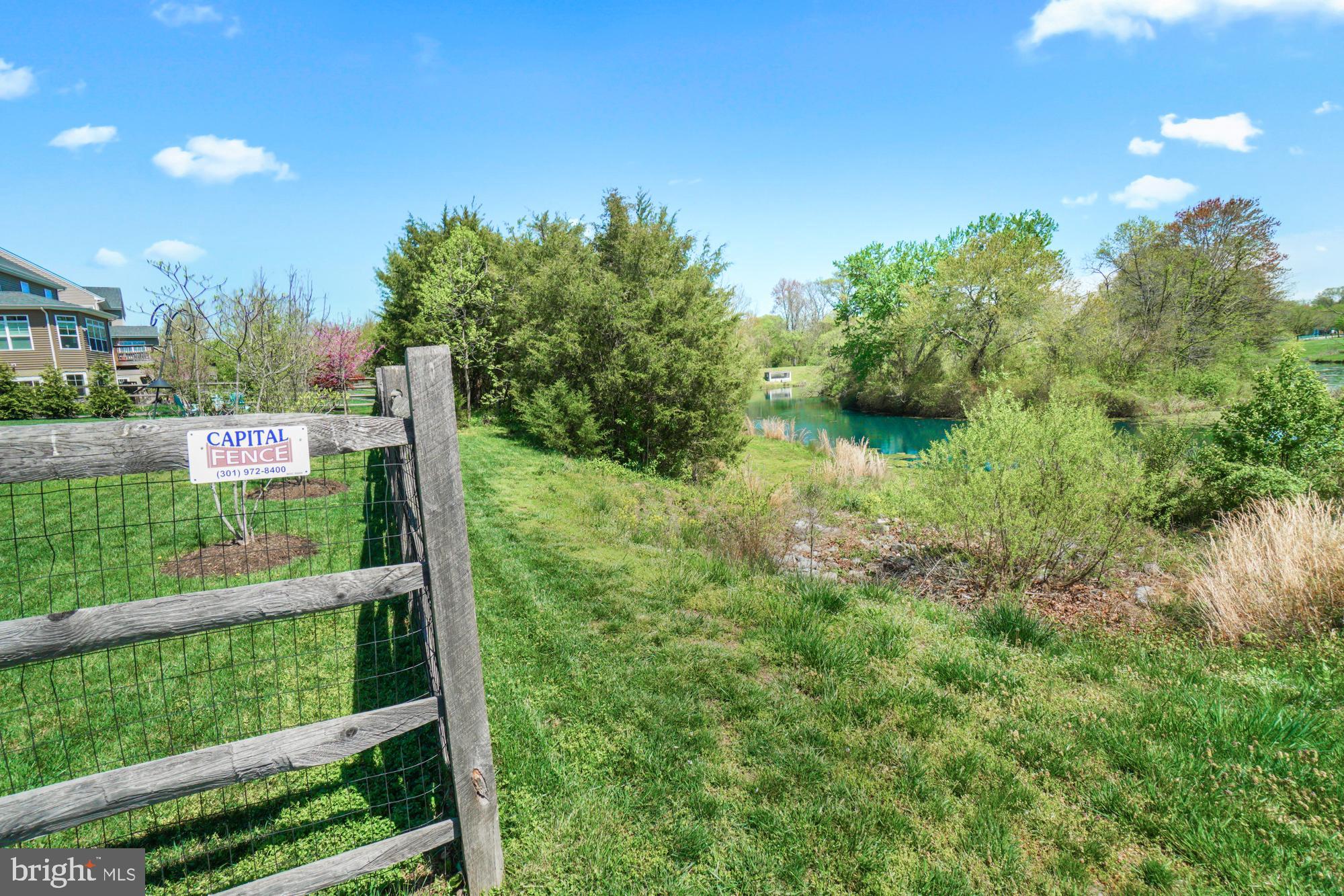 Grassy yard beside a tree-lined pond, viewed past a wooden fence.