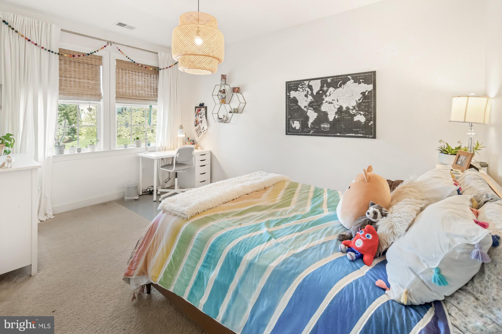 Bedroom with colorful striped bedding, plush toys, and a desk beside two windows.