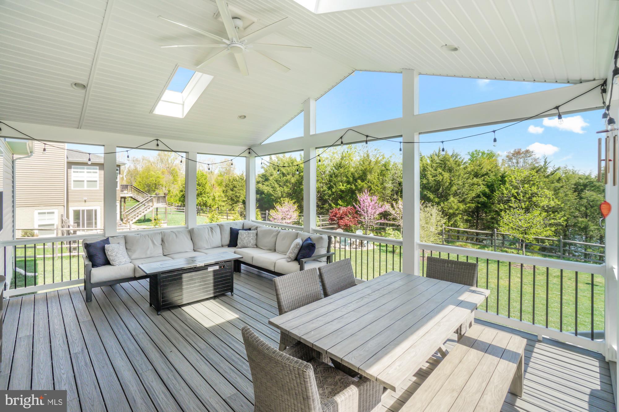 Covered deck with a sectional sofa, dining table, ceiling fan, and views of a leafy yard.