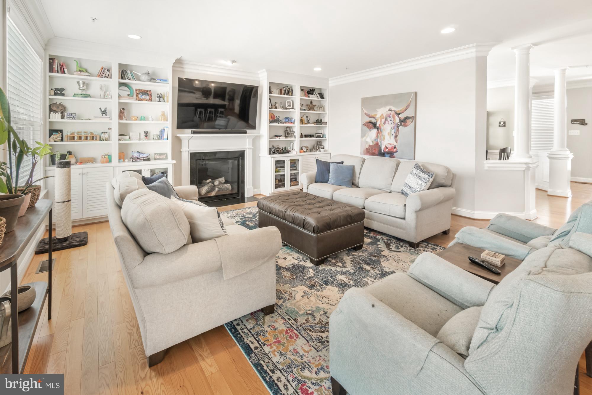 Furnished living room with built-in shelves, a fireplace, wall-mounted TV, and pale sofas around a leather ottoman.