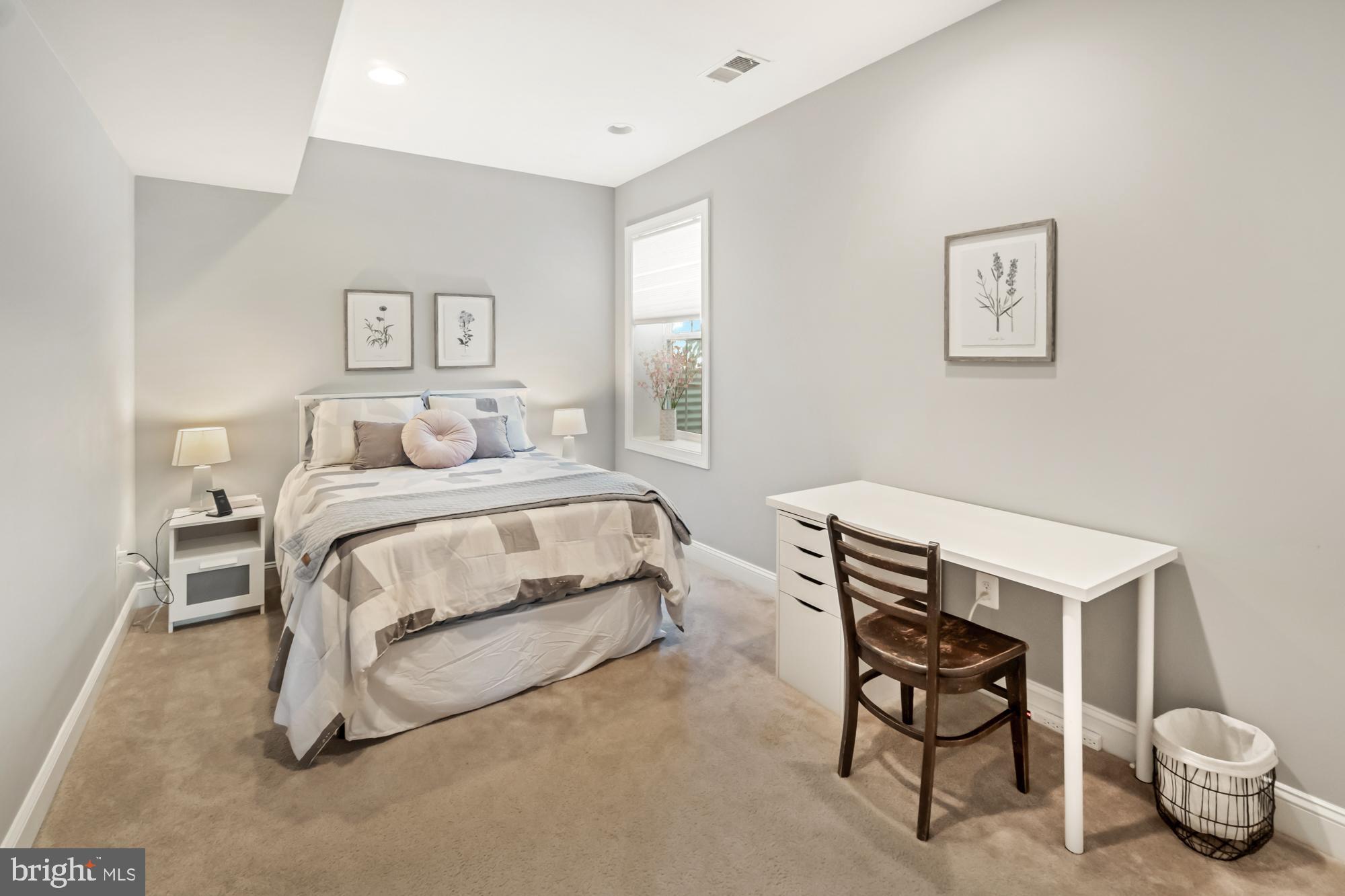 Furnished bedroom with a neutral bed, botanical prints, and a white desk and wooden chair.