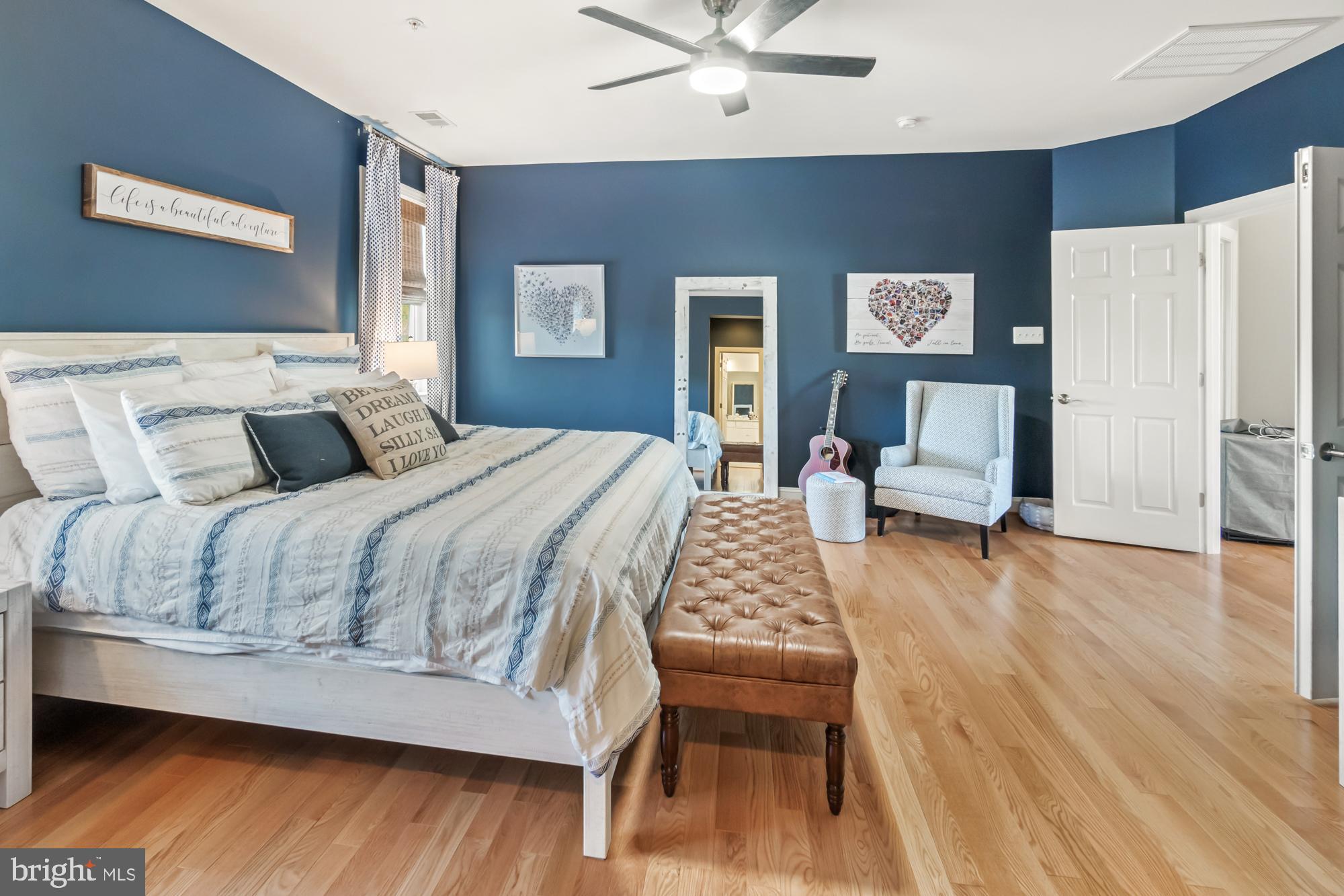 Staged bedroom with blue walls, a white bed, leather bench, hardwood floors, and a guitar beside an upholstered chair.