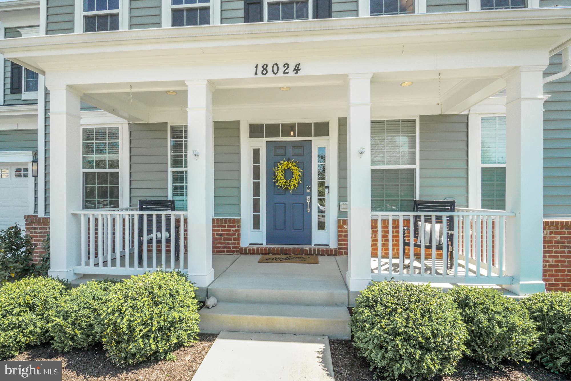 Home entrance with a covered porch, blue front door, yellow wreath, white railings, and trimmed shrubs.