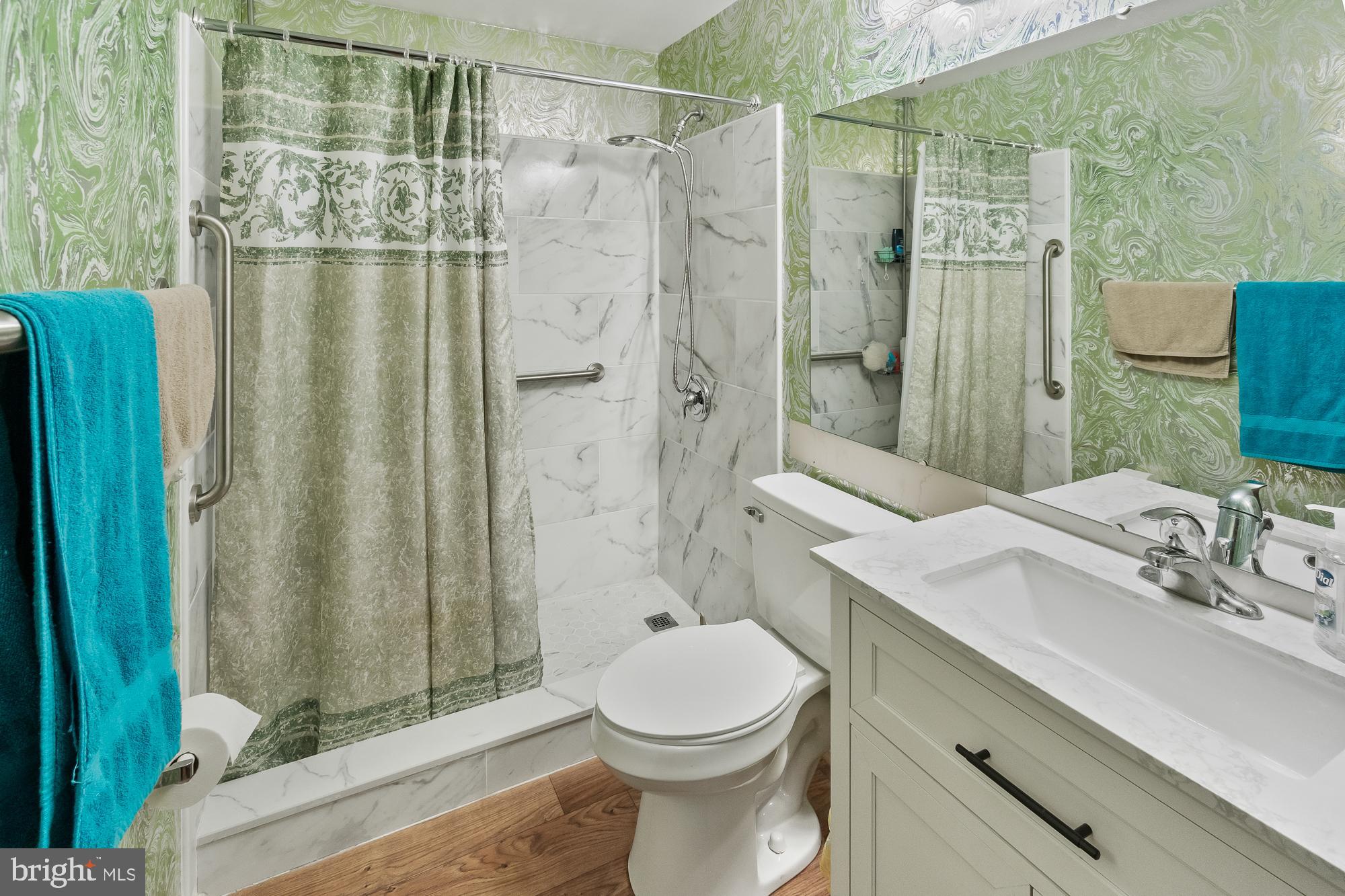 Bathroom with marble-look shower tile, green patterned wallpaper, and grab bars beside the shower and toilet.