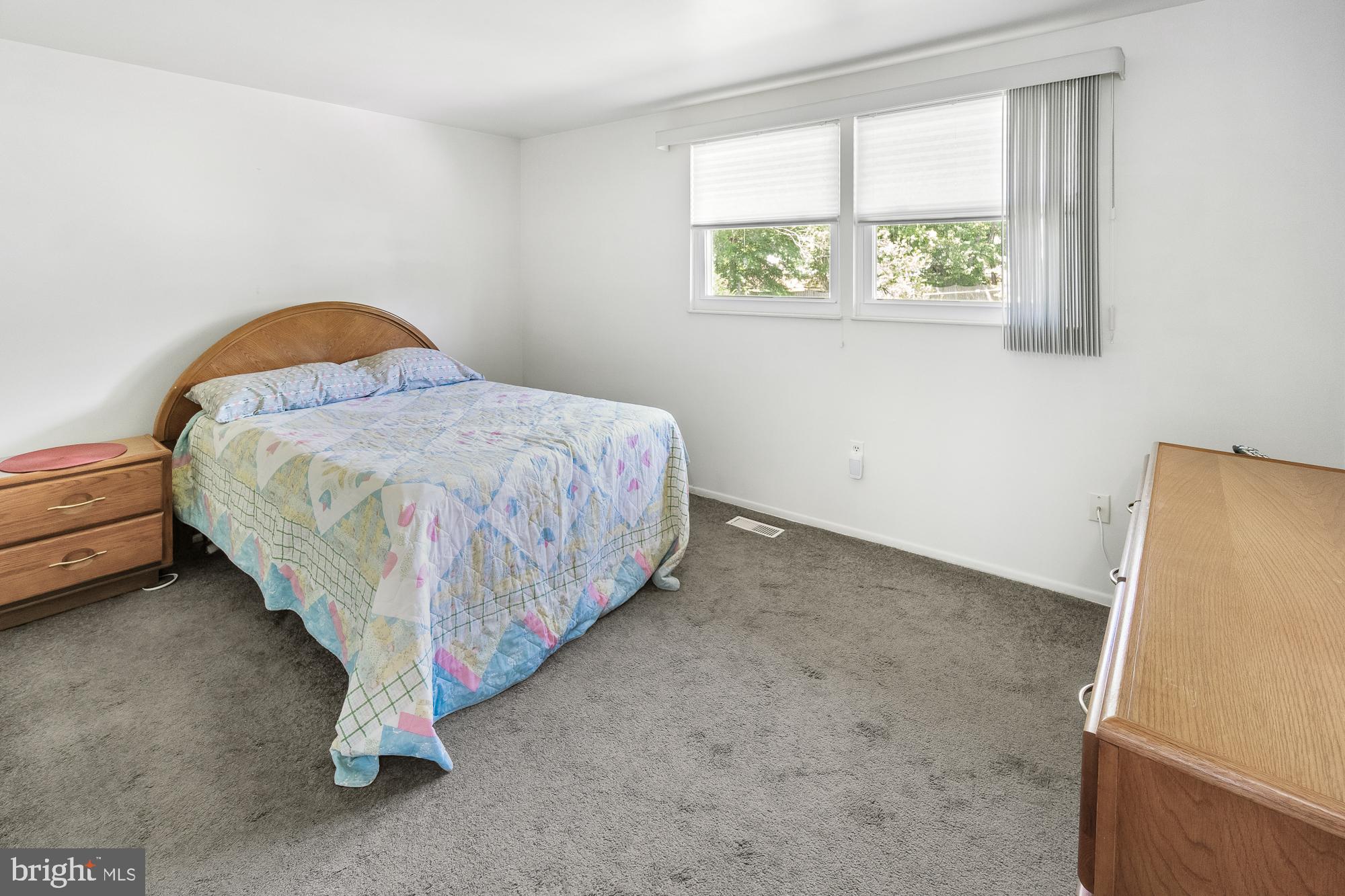 Bedroom with a wooden bed, pastel quilt, gray carpet, and two windows with blinds.