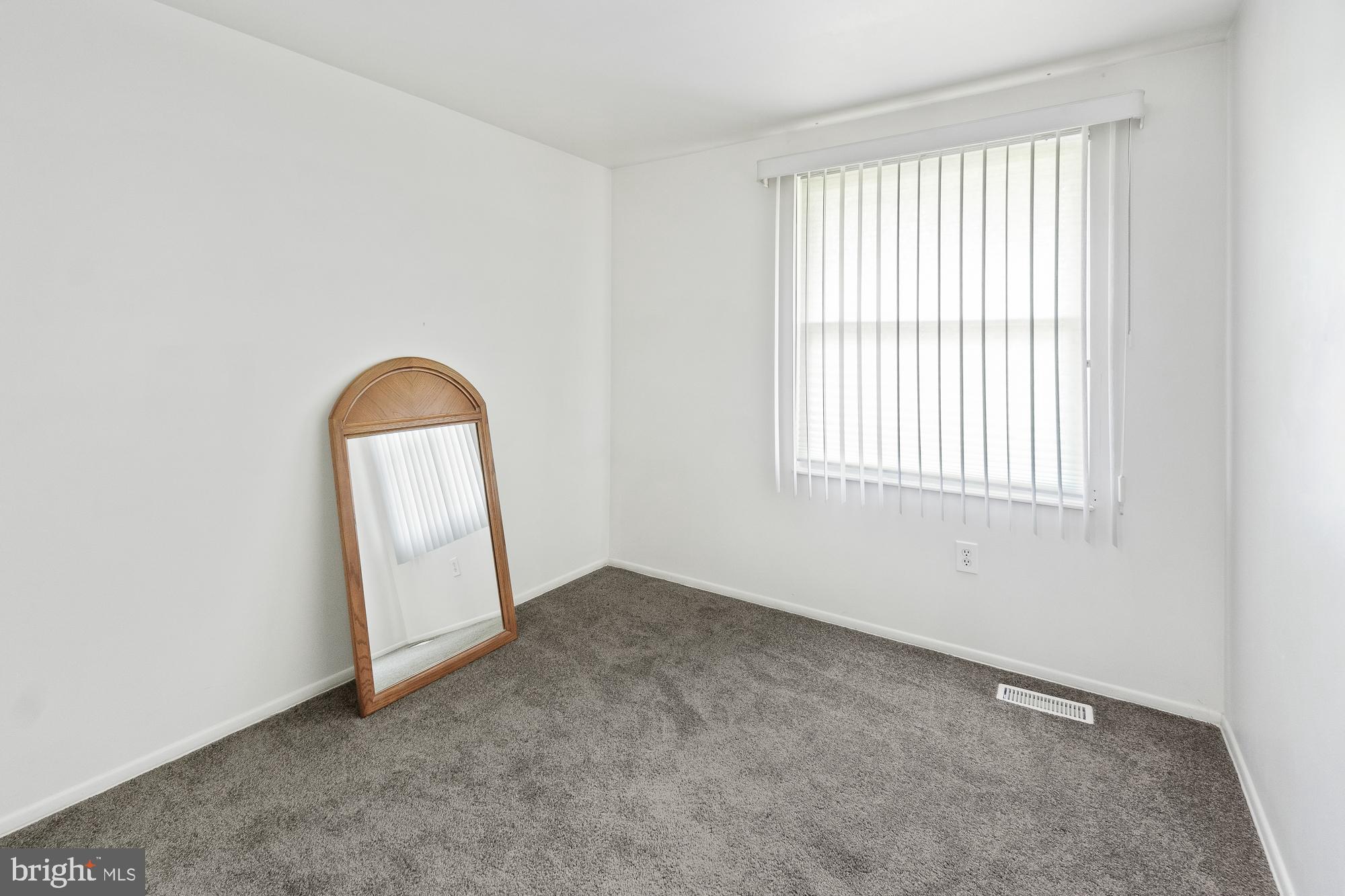 Empty carpeted bedroom with a large window and a wooden-framed mirror leaning against the wall.